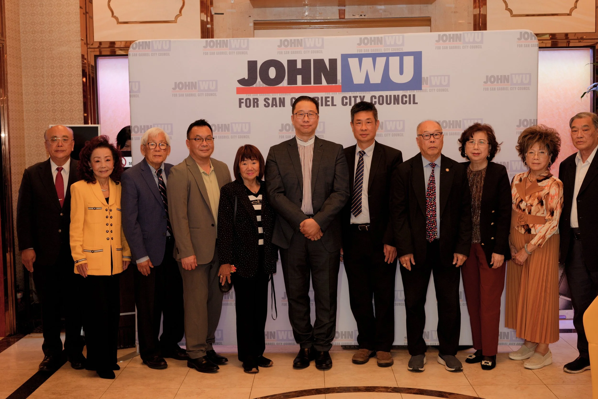 A group of eleven people standing in front of a backdrop that reads "John Wu for San Gabriel City Council." The group consists of five women and six men dressed in formal attire, posing for a photo at an indoor event.