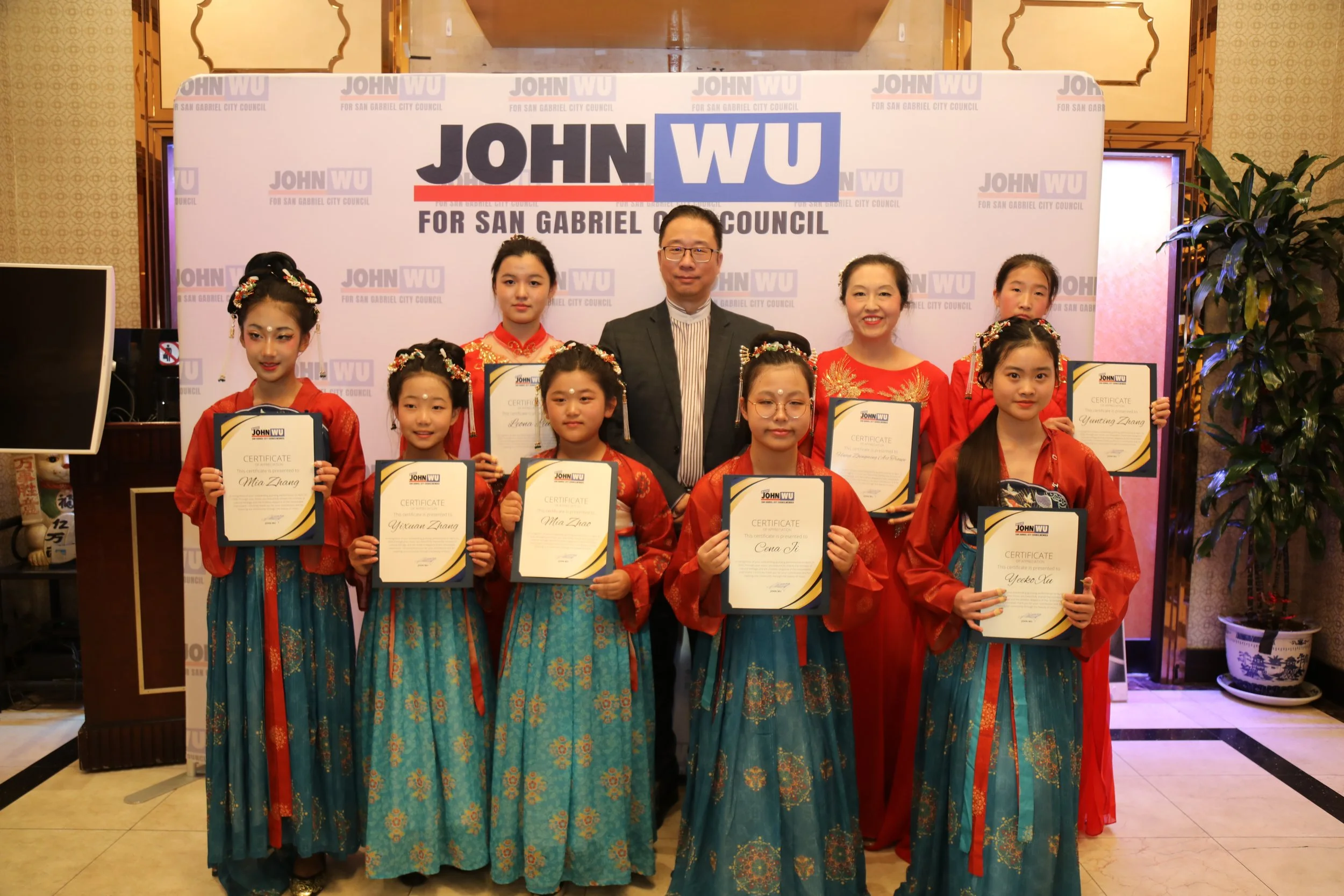 Group of nine people, including children and adults, standing in front of a backdrop with 'JOHN WU FOR SAN GABRIEL CITY COUNCIL' written on it. Children are holding certificates, dressed in traditional red and blue cultural attire, with some wearing 