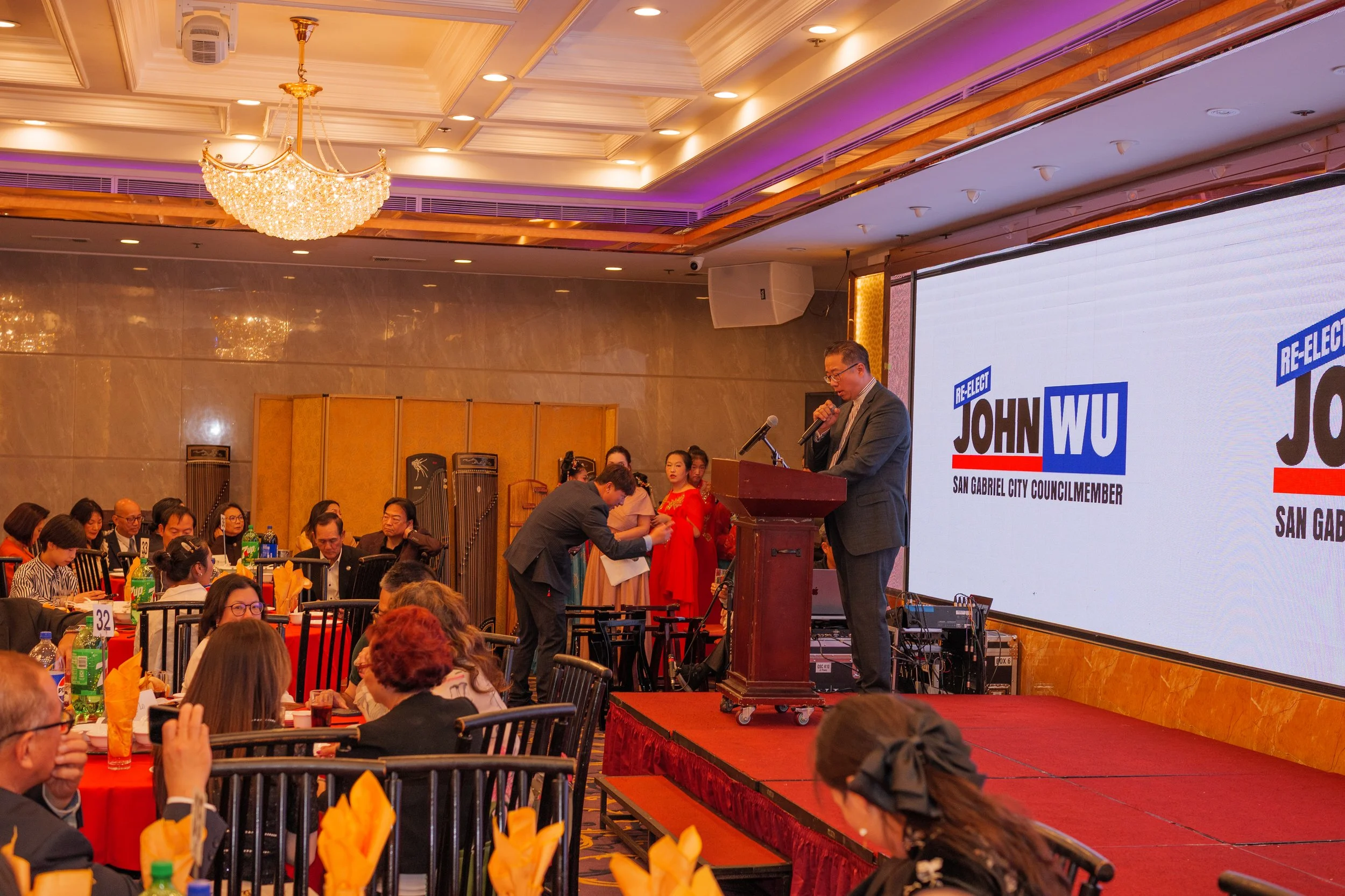 A man in a suit and glasses speaking at a podium during a political event with a large screen behind him displaying 'RE-ELECT JOHN WU, SAN GABRIEL CITY COUNCILMEMBER'. The audience is sitting at tables with orange napkins, bottles of soda, and food, 