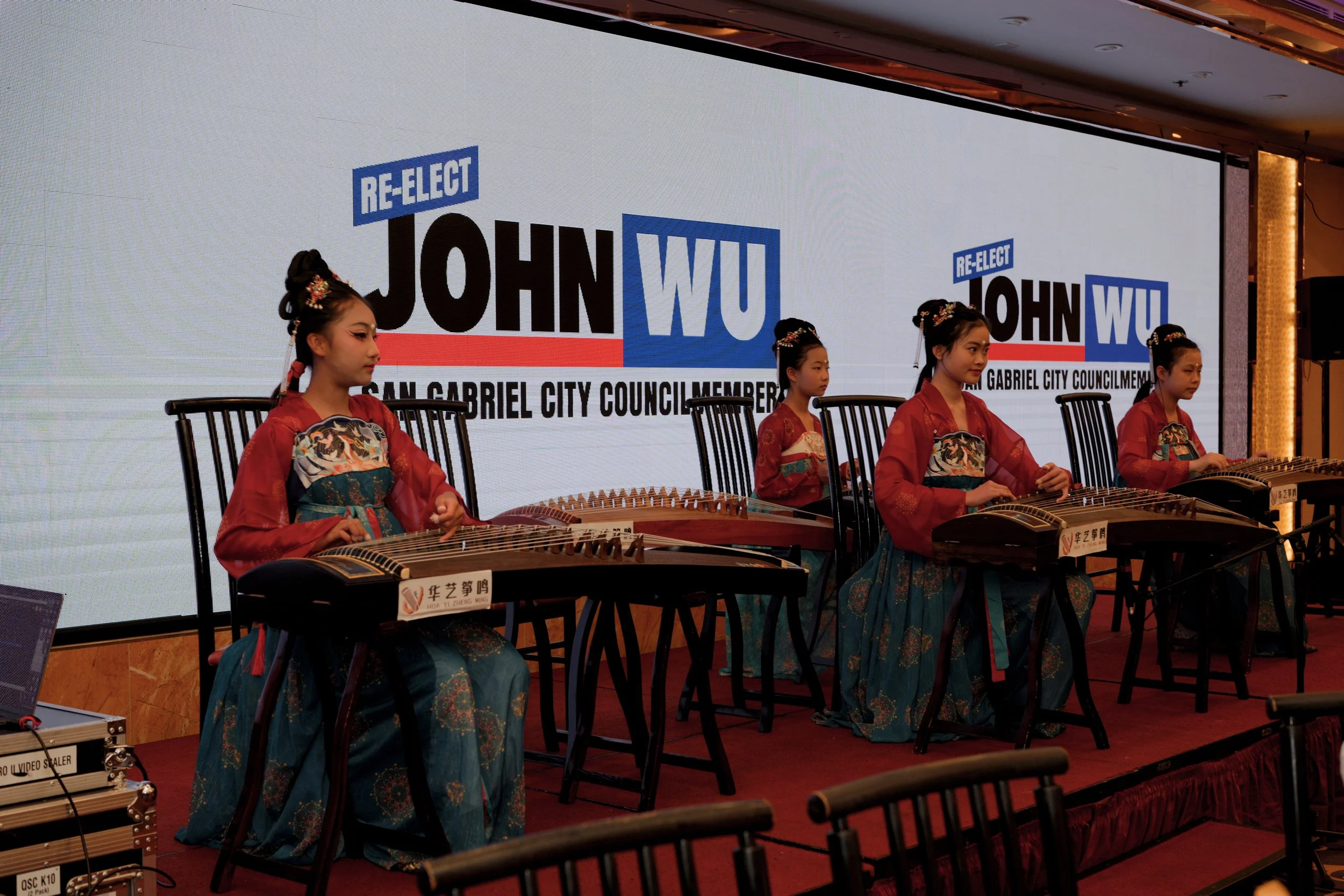 Young women in traditional Asian attire playing musical instruments on stage during a political event, with a large screen behind them displaying campaign slogans for John Wu re-election for San Gabriel City Council.