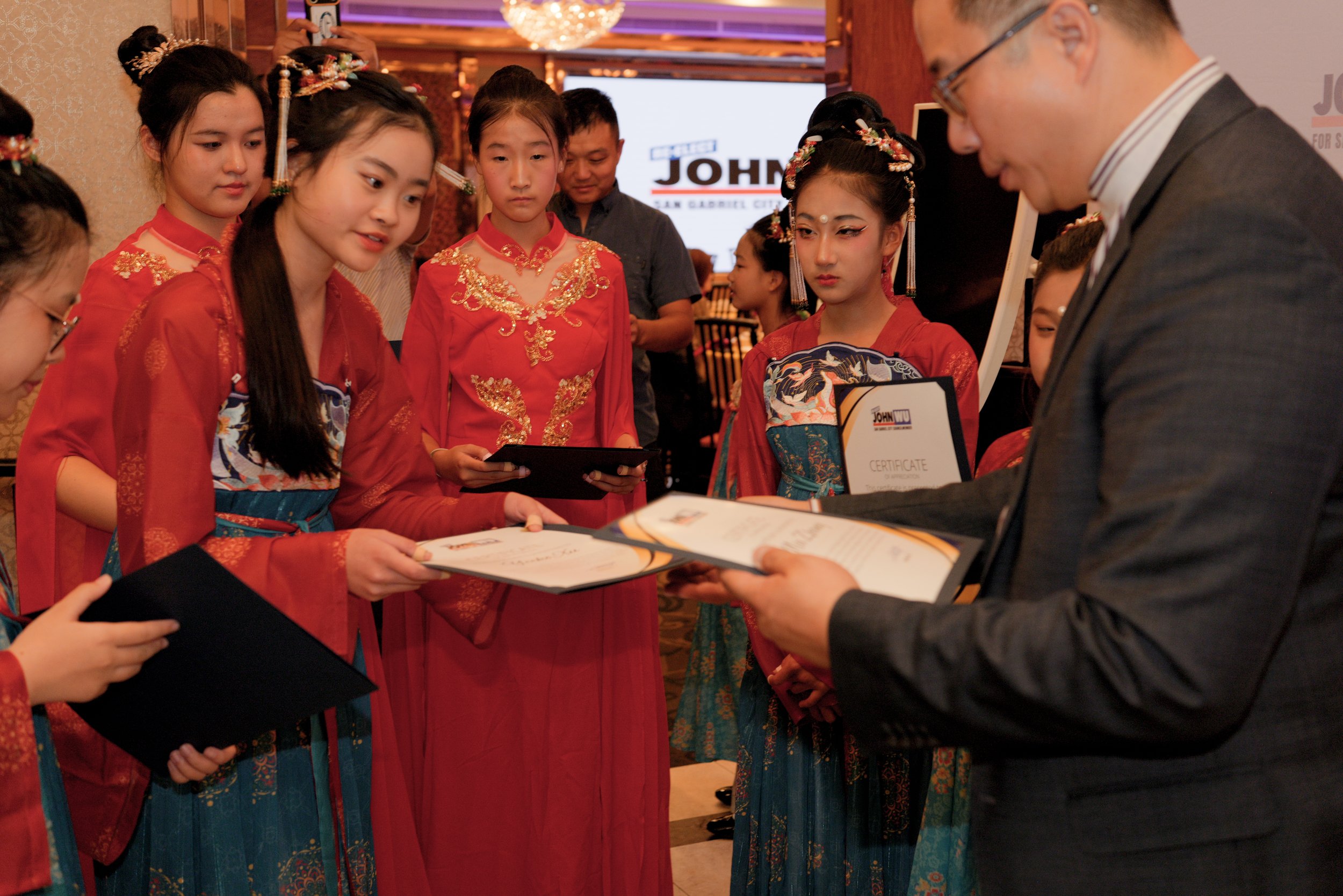 Group of young women dressed in traditional Asian attire receiving certificates from a man in a suit at an indoor event.