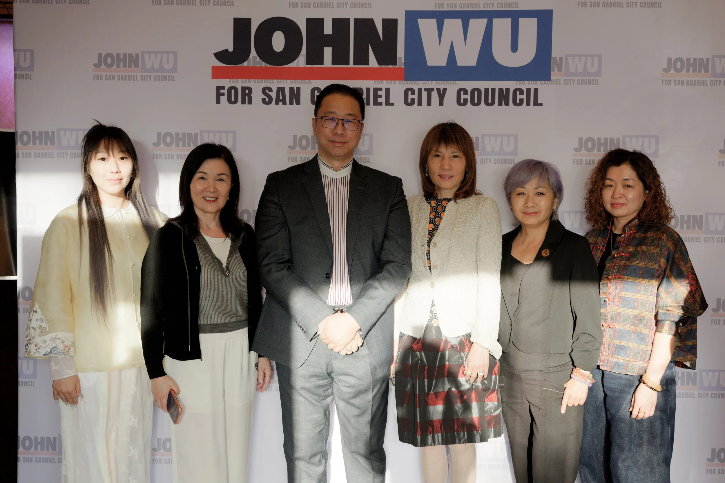 Group of six people standing in front of a backdrop with text supporting John Wu's candidacy for San Gabriel City Council. The group includes five women and one man, all dressed in formal attire.