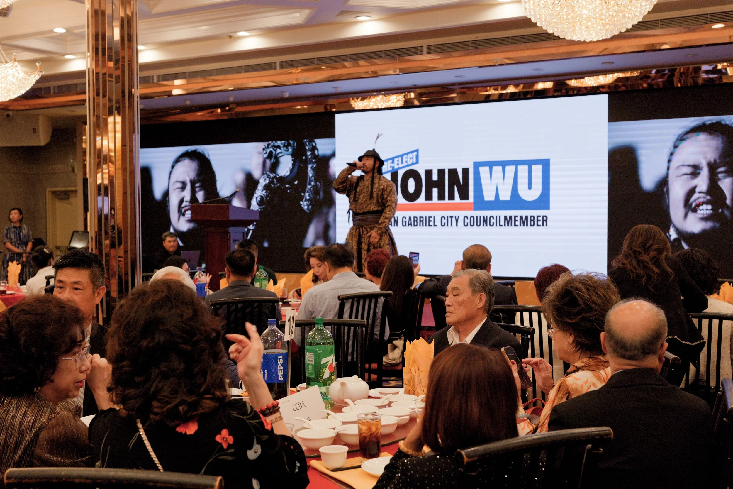 A man dressed in a traditional outfit is speaking on stage at a formal event, with a large screen behind him displaying his image and name, 'John Wu,' a candidate for city council in San Gabriel. The audience is seated at round tables in a banquet ha