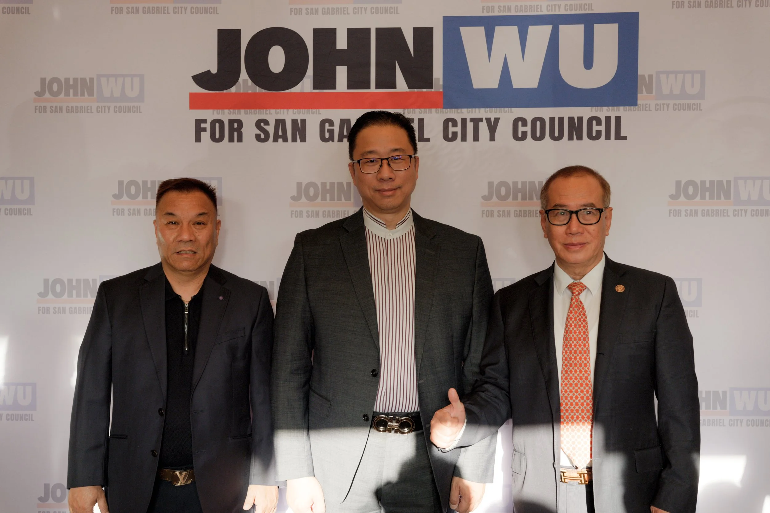 Three men standing in front of a backdrop with the 'John Wu for San Gabriel City Council' logo. The man in the middle is wearing glasses, a dark suit, and a striped shirt, and is giving a thumbs-up. The man on the left is wearing a black jacket, and 
