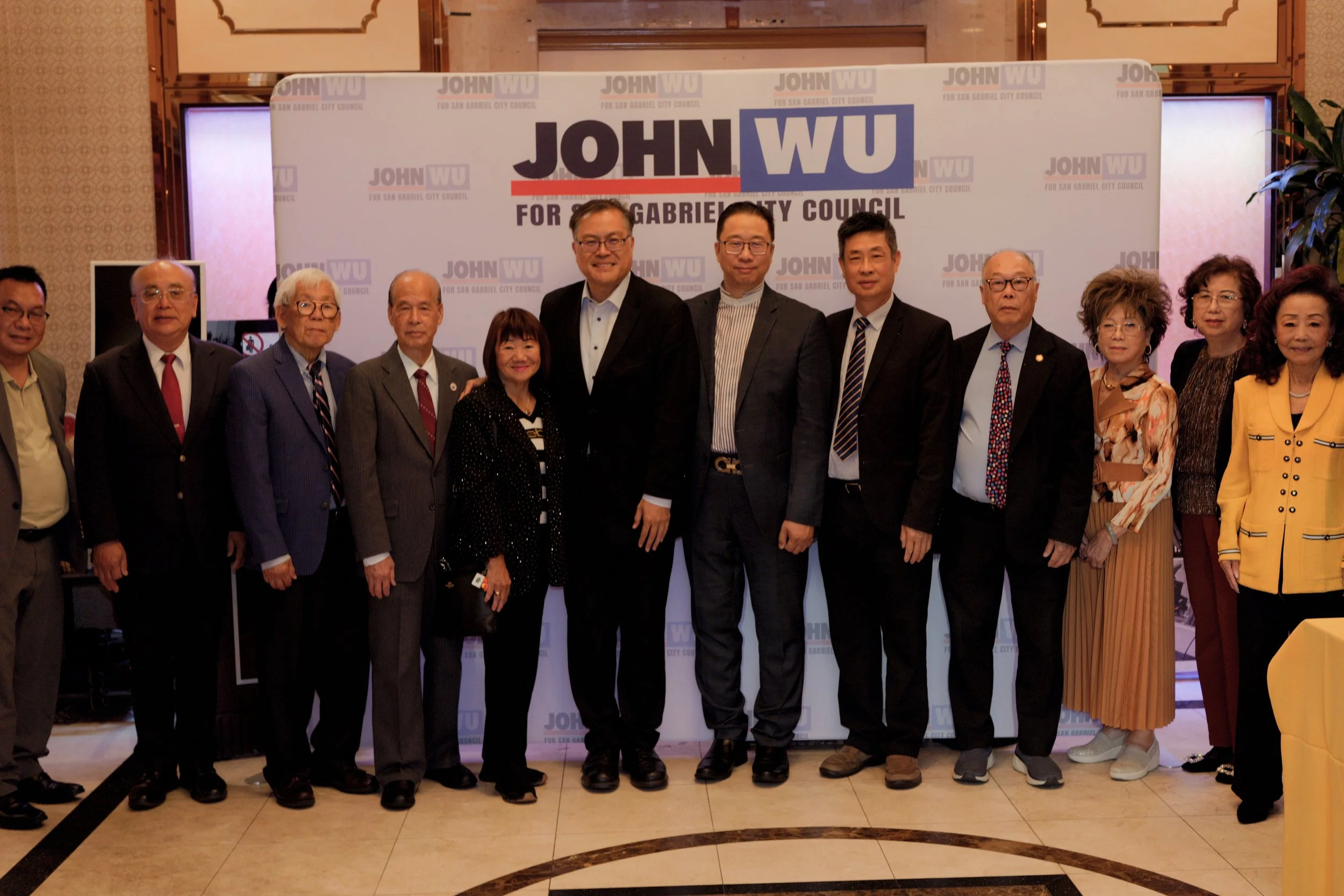 A group of people standing in front of a campaign sign that reads 'John WU for San Gabriel City Council' at an indoor event.