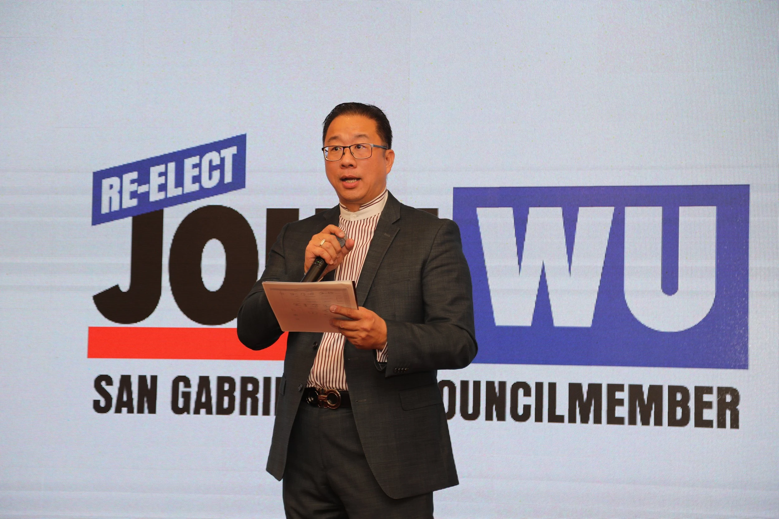 A man wearing glasses and a suit speaking into a microphone at a political event, with a large screen behind him displaying the words 'Re-Elect' and 'San Gabriel Youth Leader'.