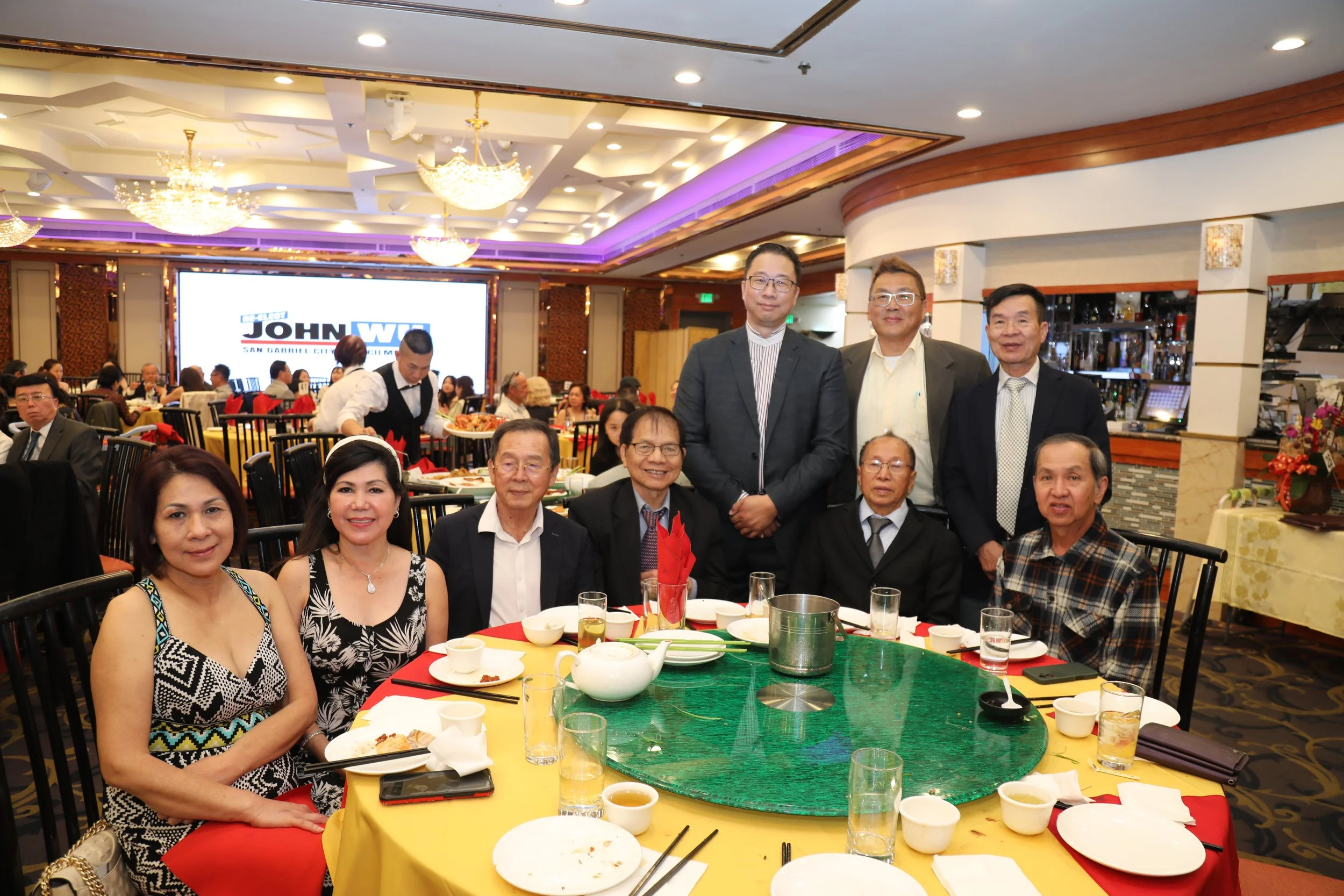 A group of people gathered around a restaurant table with food, drinks, and utensils, posing for a photo, with a large screen and other diners in the background.