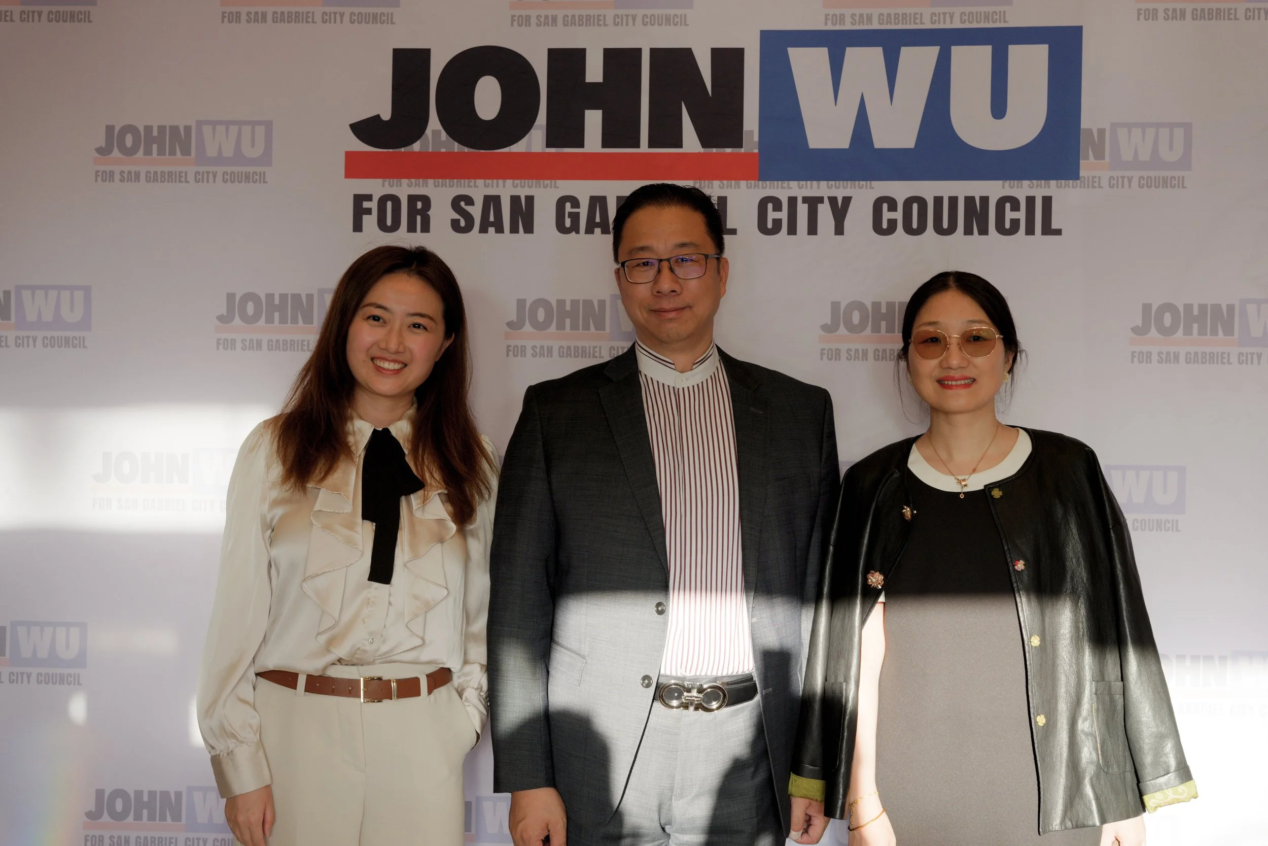 Three people standing in front of a campaign backdrop for John Wu, a candidate for San Gabriel City Council. Two women and one man, dressed in business casual and semi-formal attire, smiling at the camera.