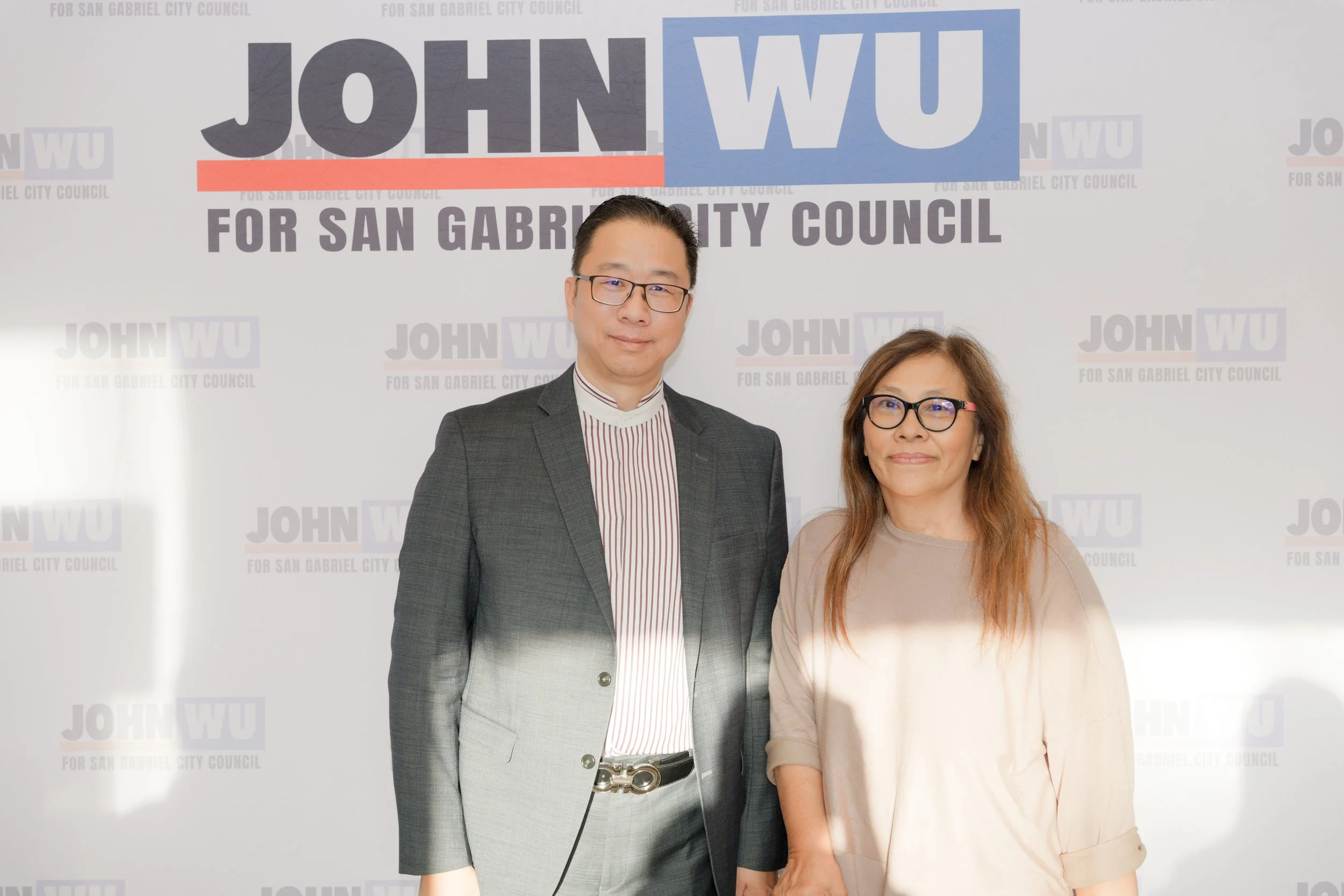 A man and a woman stand in front of a backdrop with text that reads 'JOHNWU FOR SAN GABRIEL CITY COUNCIL'. The man is wearing glasses, a dark plaid suit, and a striped shirt, and the woman has glasses and wears a beige top.
