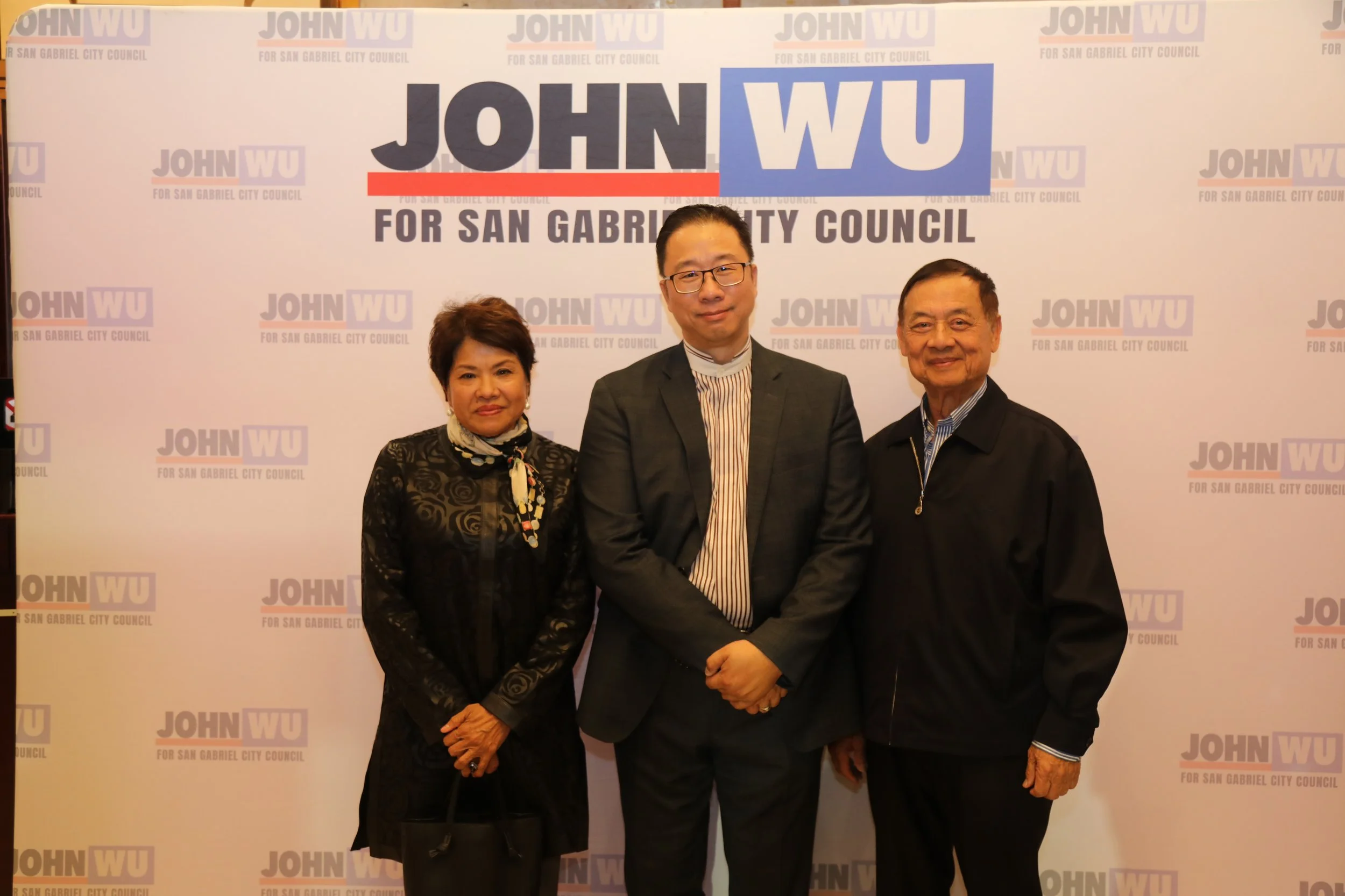 Three people standing in front of a campaign banner for John Wu for San Gabriel City Council. Two men and one woman, all dressed in formal attire, smiling at the camera.