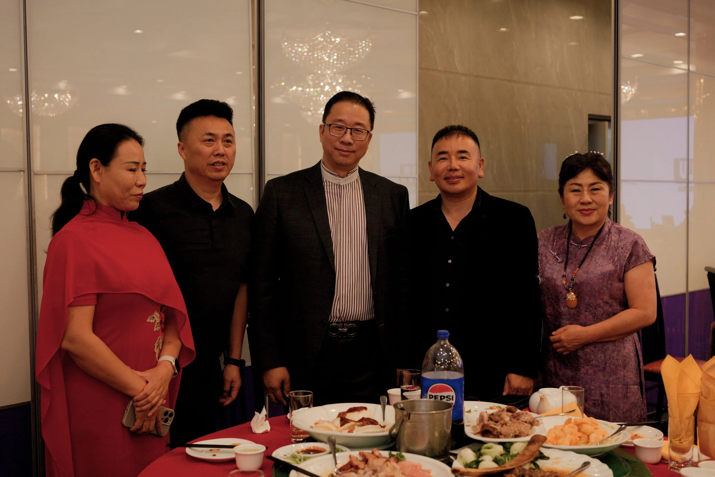 A group of five people standing behind a table of food at a formal event. The table has various dishes, including meats and vegetables. The background features a chandelier and a reflective wall.
