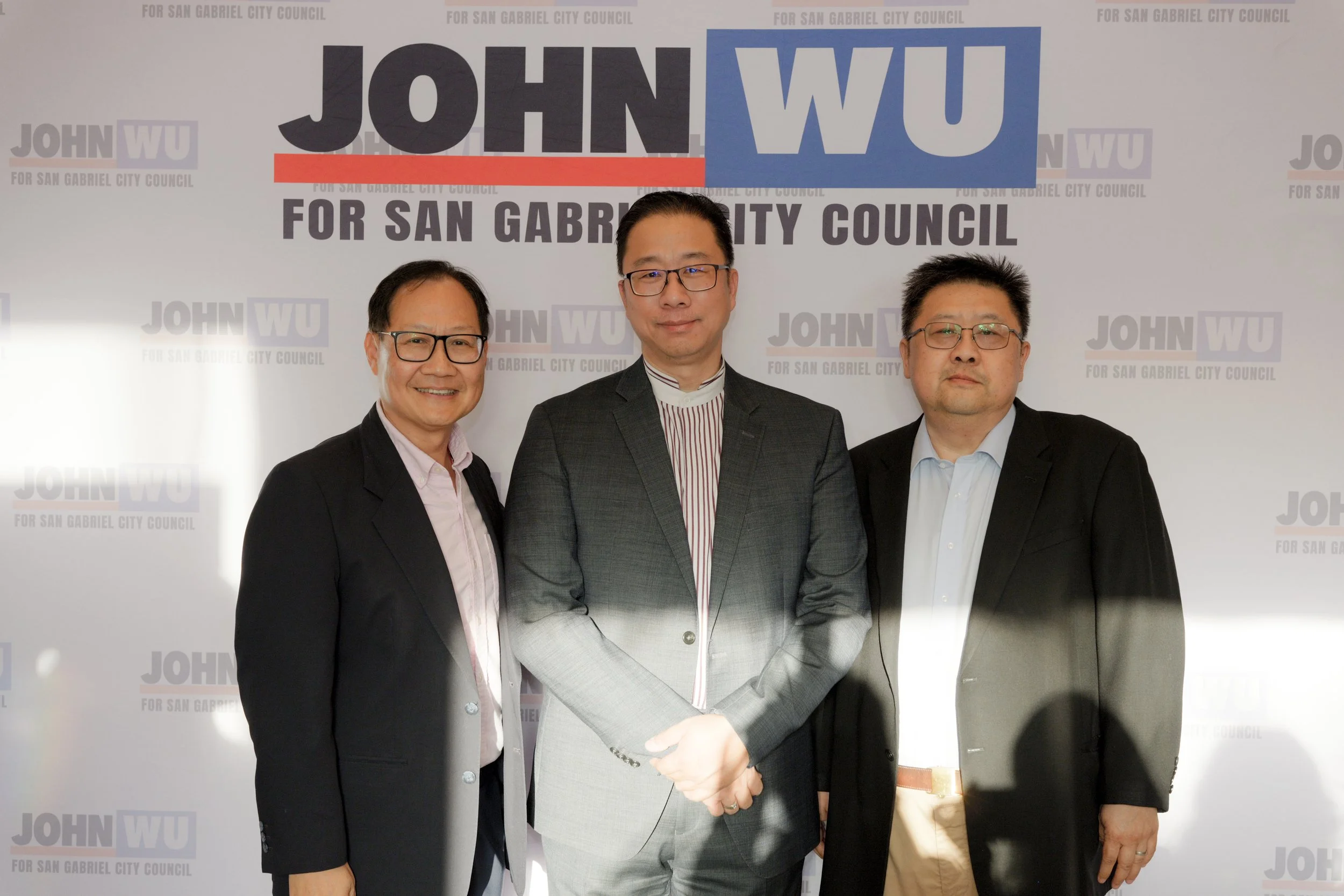 Three men in suits standing in front of a backdrop with the text 'JOHN WU for San Gabriel City Council' printed repeatedly. The man in the middle is shaking hands with someone off-camera.