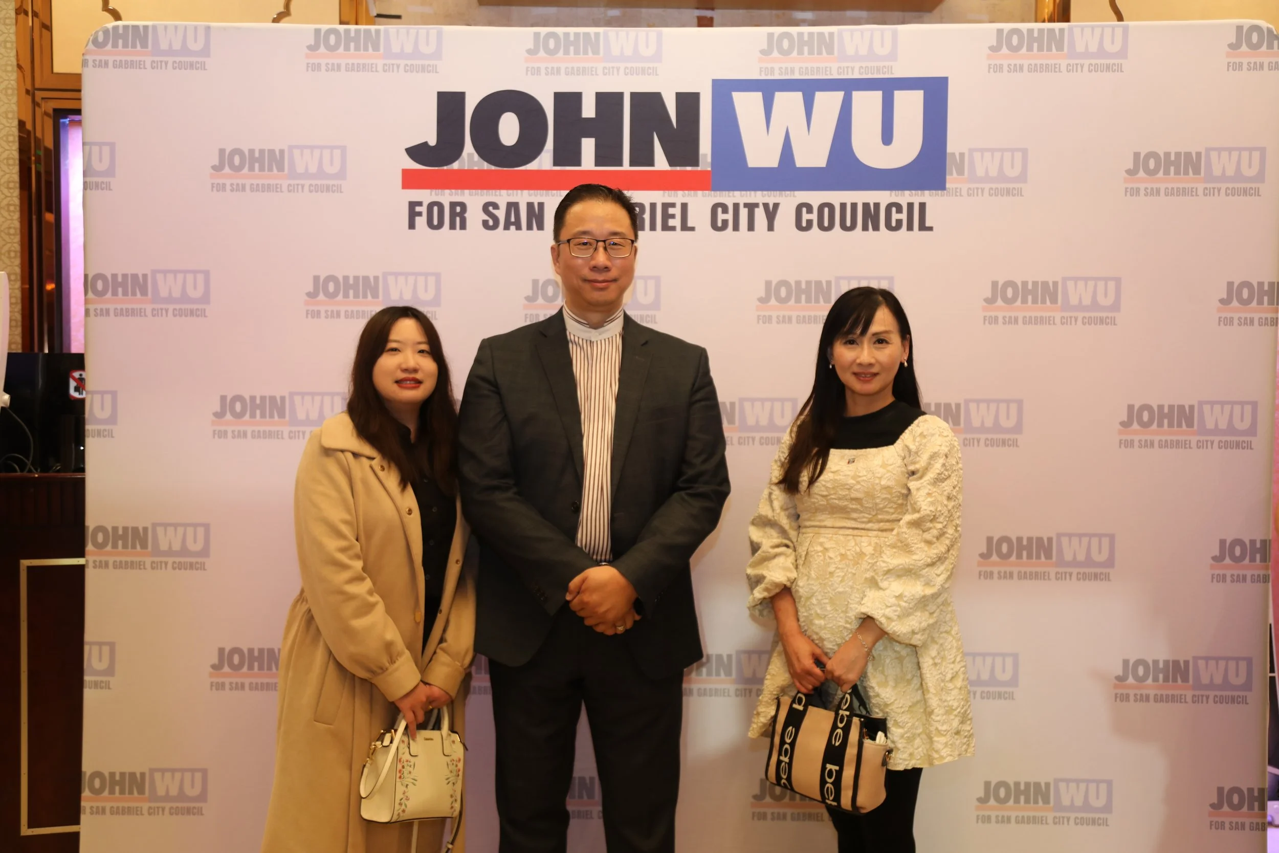 A group of three people standing in front of a white backdrop with the text "John WU for San Gabriel City Council" repeated in purple, pink, and black. The person in the middle is a man wearing glasses and a suit, flanked by two women, one in a beige