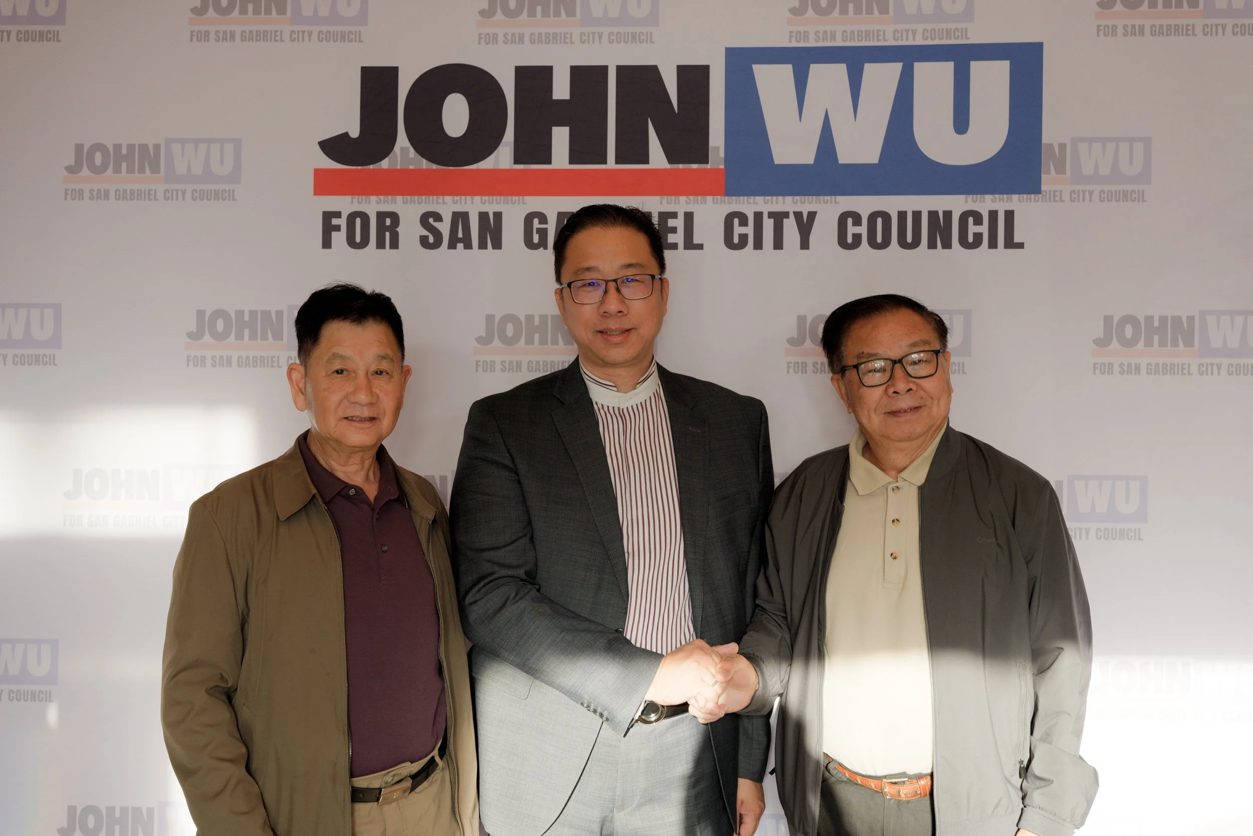 Three men are standing together during a handshake, with a background sign that says 'JOHN WU for San Gabriel City Council.'