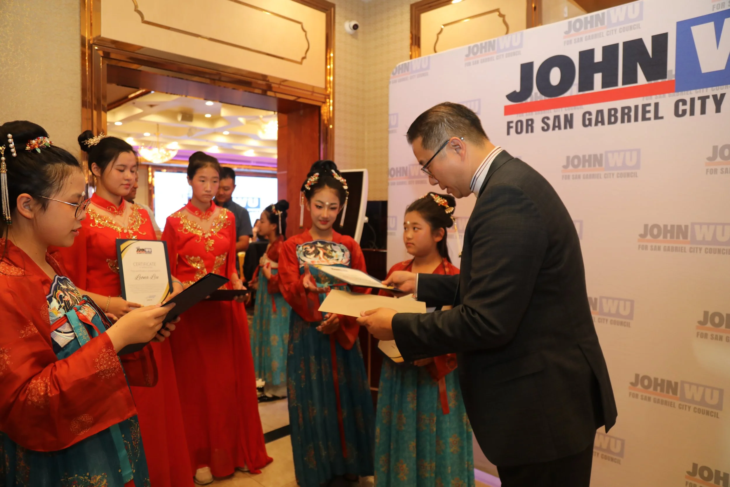 A man in a suit receiving a certificate from a young girl dressed in traditional Chinese attire, with other girls in similar attire standing beside him during an award ceremony.