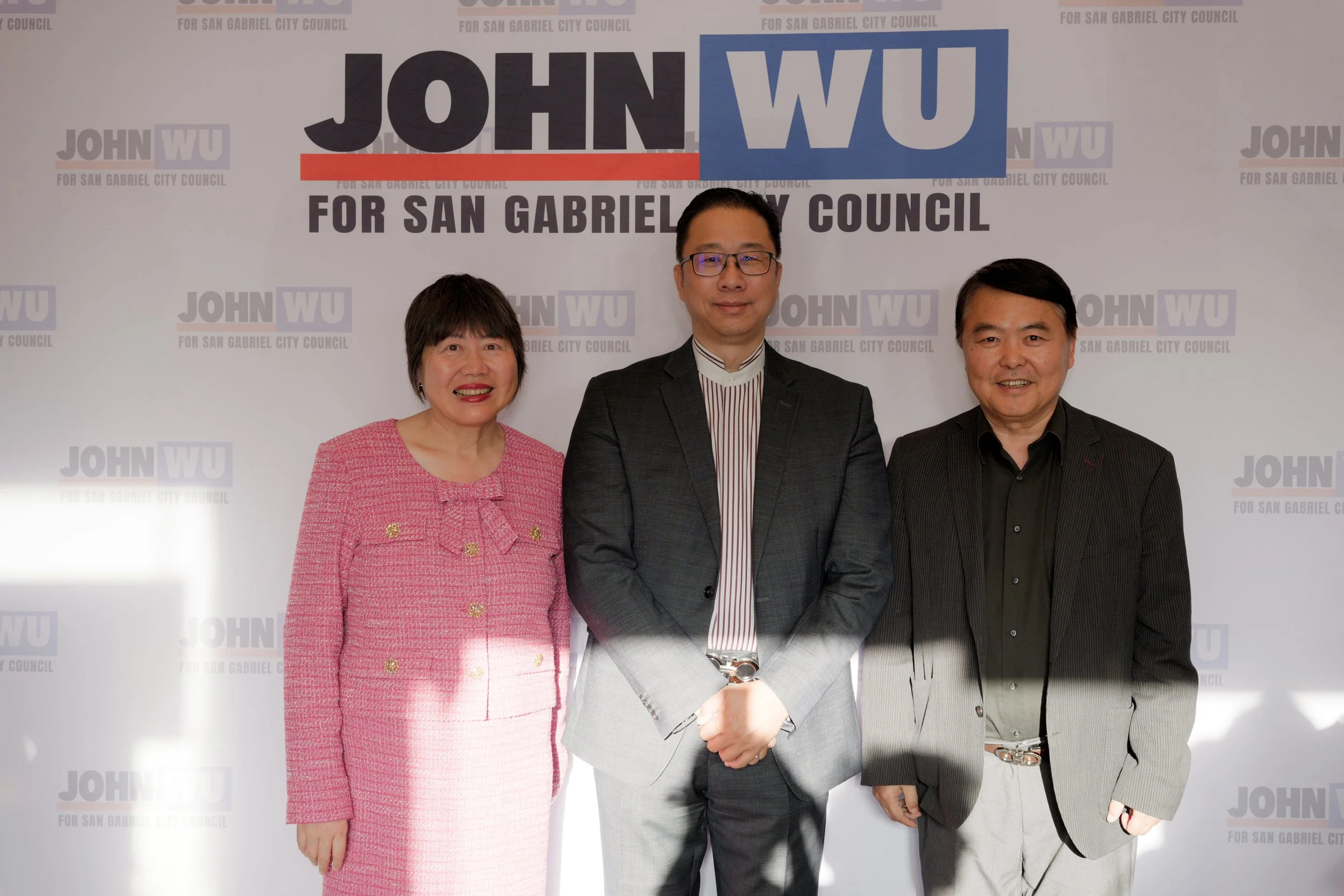 Three people standing in front of a campaign background. The background has the text 'John Wu for San Gabriel City Council' repeated across it. The person on the left is a woman wearing a pink blazer, the person in the middle is a man with glasses in