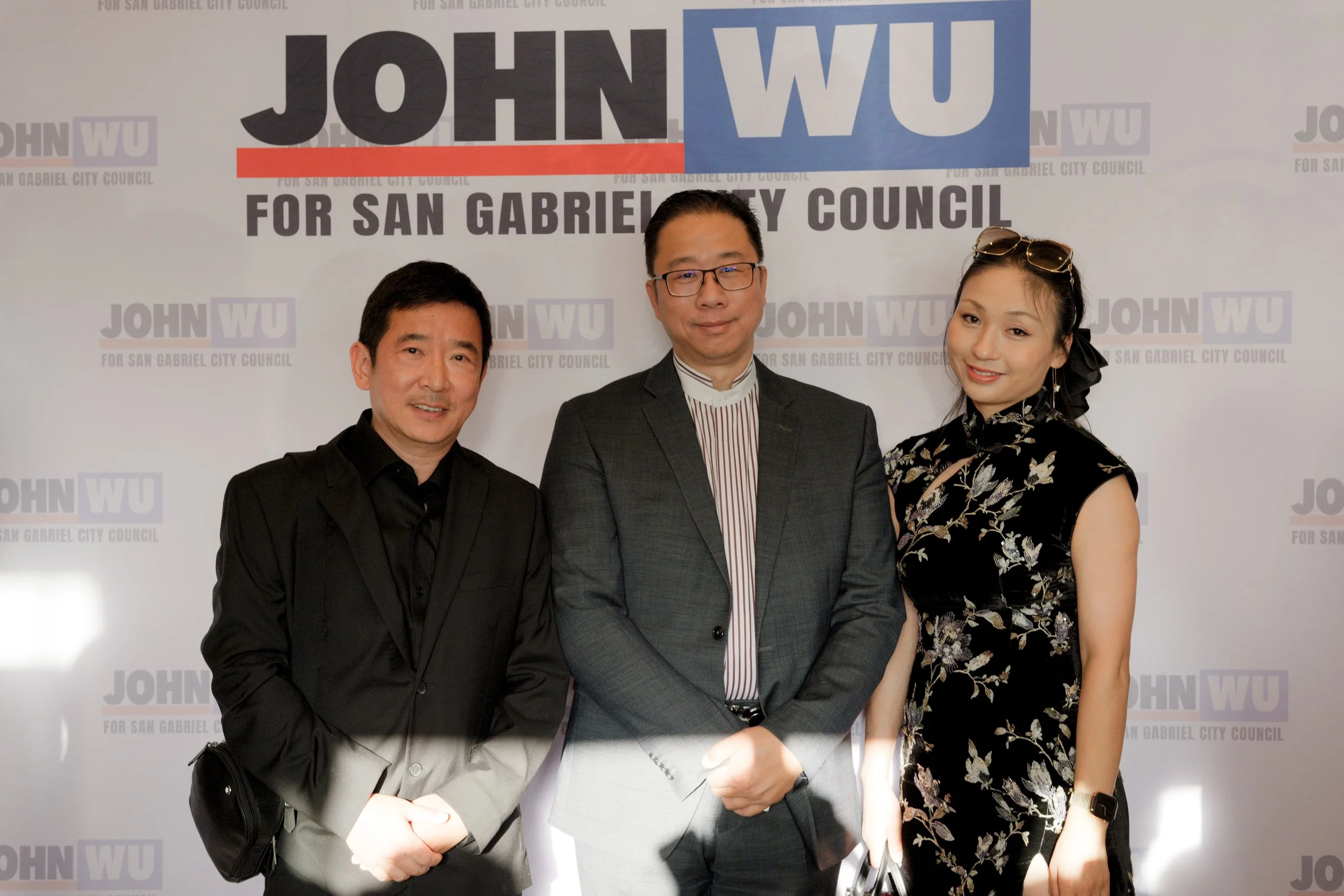 Three people posing in front of a background with the text "JOHN WU for San Gabriel City Council." The man on the left is wearing a black shirt, the man in the center is wearing glasses and a gray suit, and the woman on the right is wearing a floral 