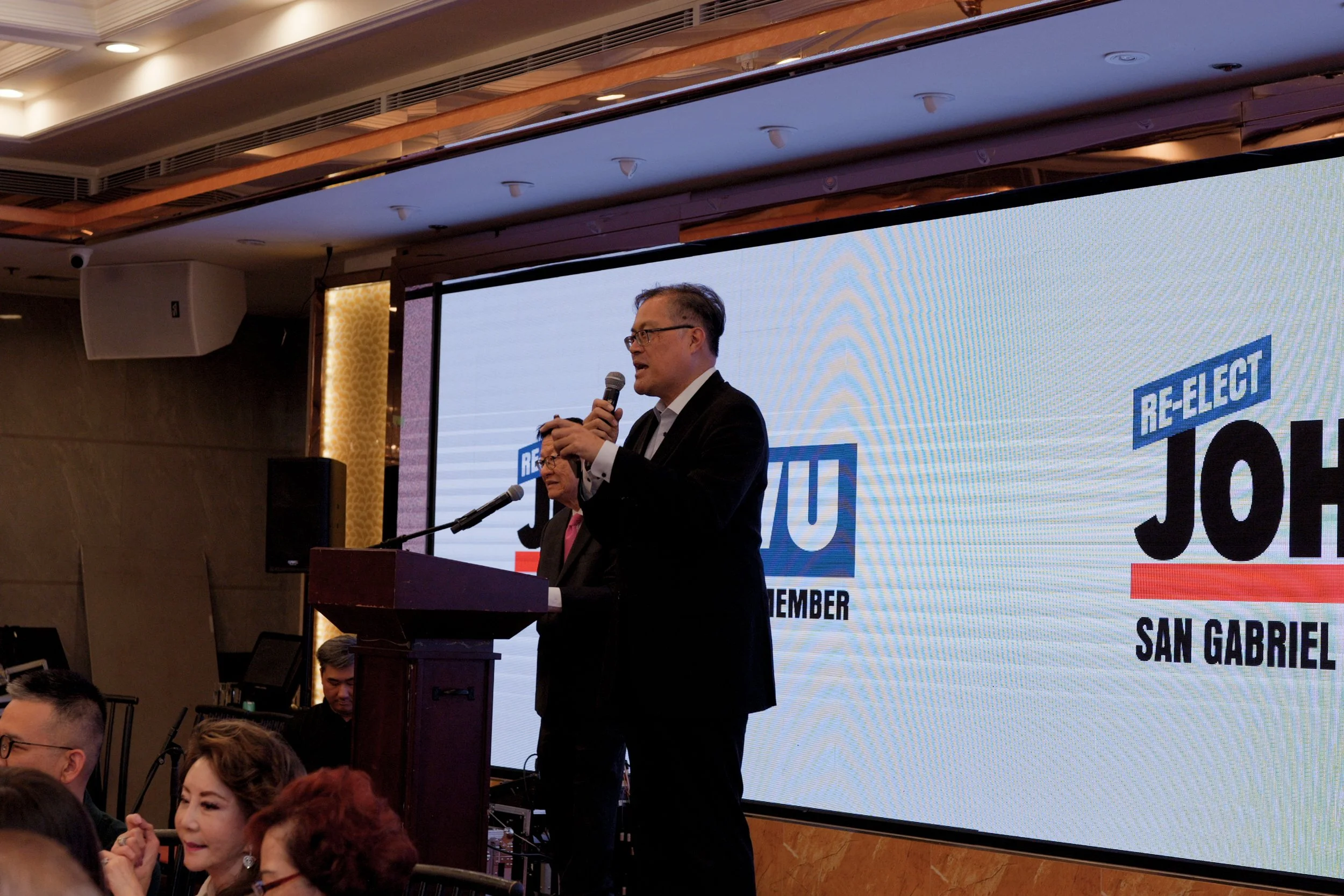 A man in a black suit and glasses speaking into a microphone on a stage in front of a large screen that reads "RE-ELECT" and "JOSH" during an event. Several audience members are visible, including a woman with red hair in the foreground.