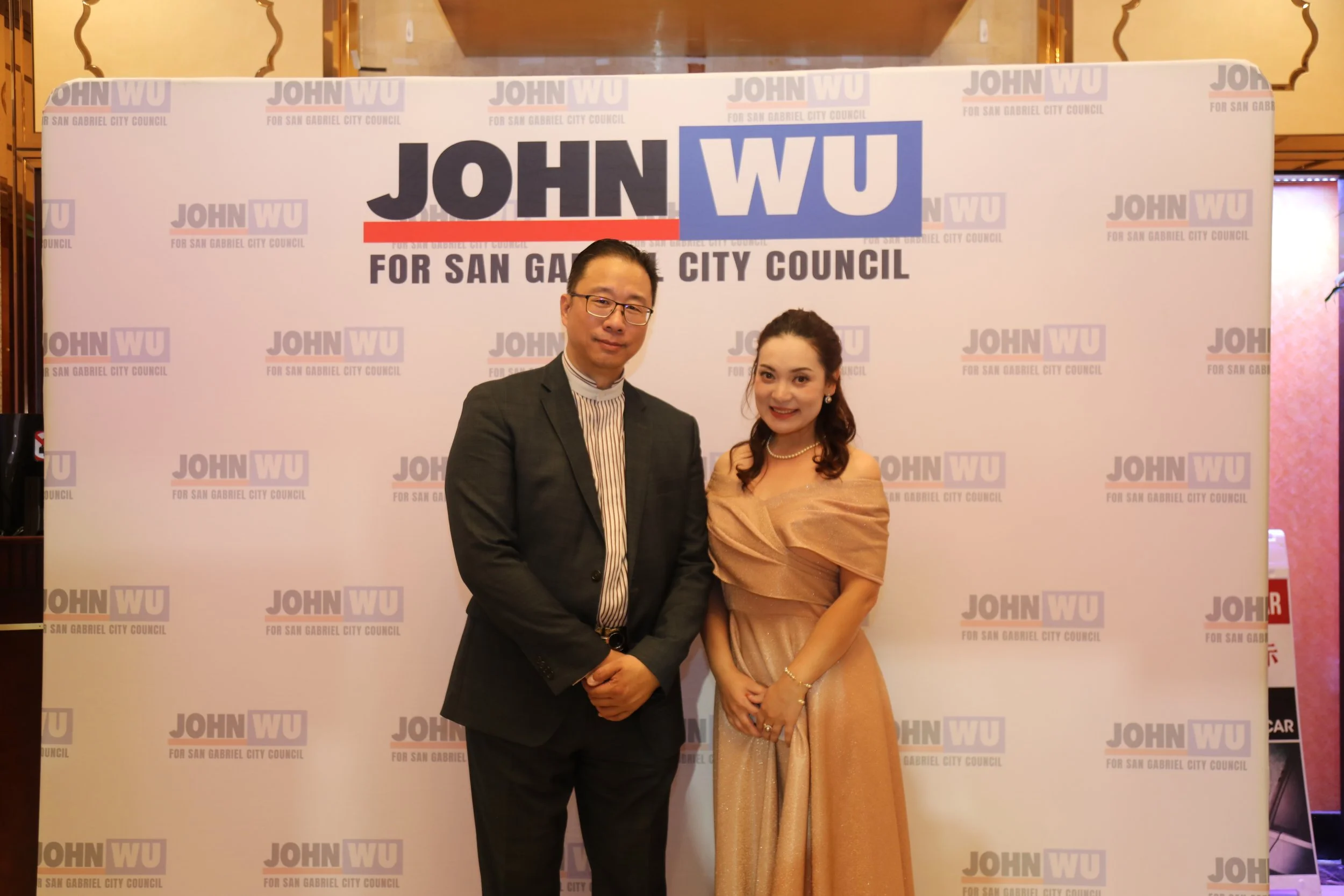 A man in a dark suit and a woman in a gold dress standing together in front of a backdrop that reads 'John W. WU for San Gabriel City Council' repeatedly. They are smiling and holding hands.
