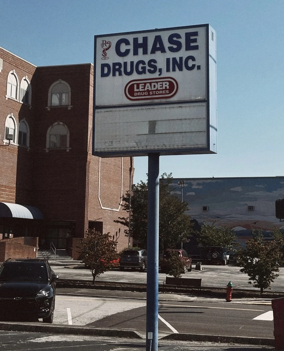 A large, rectangular sign for Chase Drugs, Inc., a pharmacy and drug store, with a red and white logo reading 'Leader Drug Stores', in a parking lot in daytime. The sign is empty below the logo. The parking lot has cars, trees, a fire hydrant, and a 
