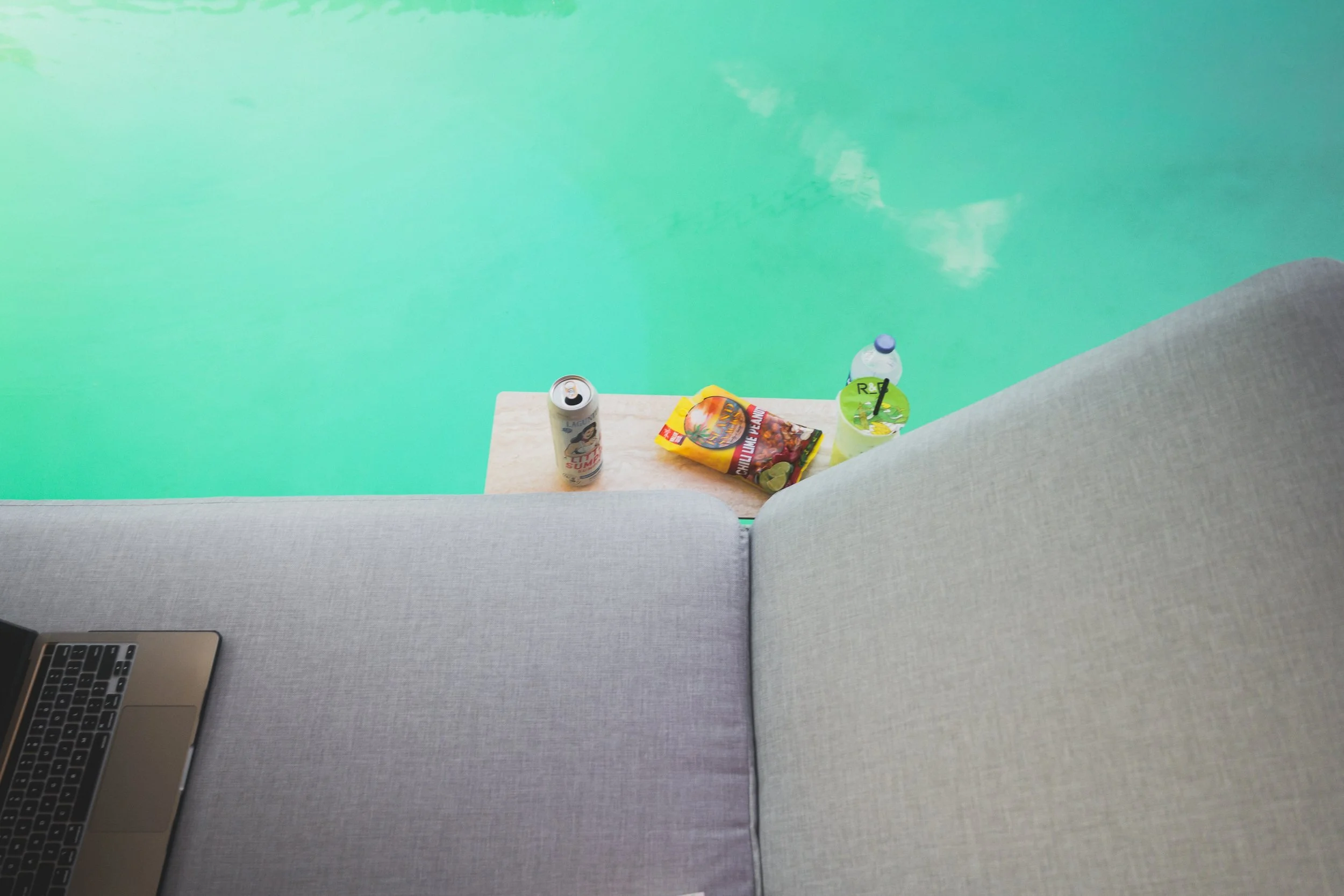 Top-down shot of the gray chaise lounge and side table with a drink can, snacks and a water bottle over turquoise pool water