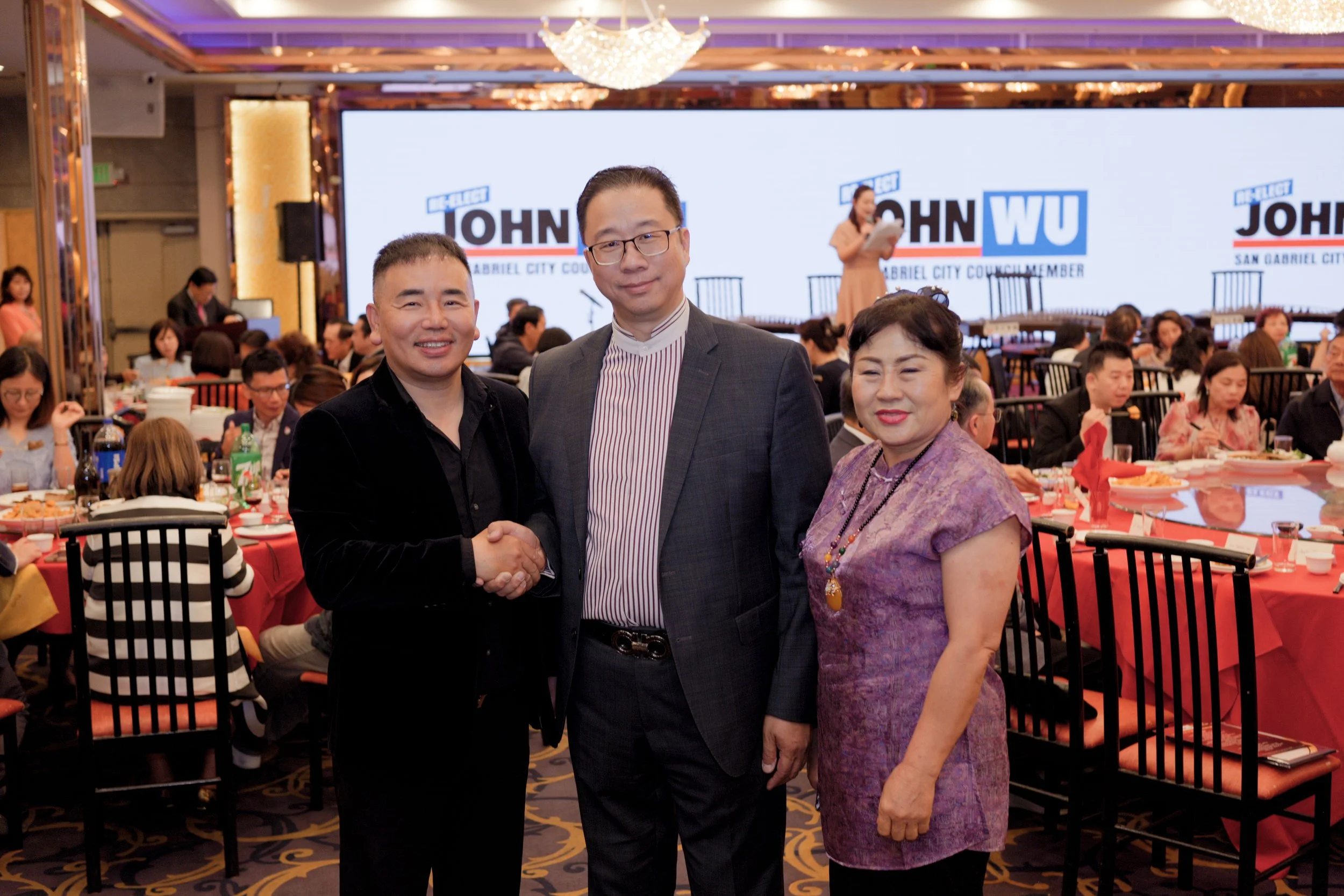 Three people standing in front of a stage with a large screen displaying campaign signs for John W. in San Gabriel City Council. The two men and one woman are smiling, with the man in the middle shaking hands with the woman.