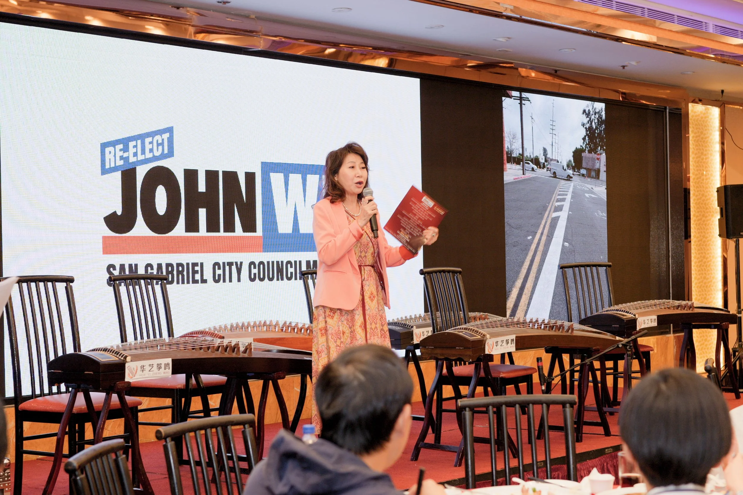 A woman in a coral blazer and dress, holding a microphone and reading from notes, standing on stage in front of a large screen displaying election campaign graphics for John W. with the text "RE-ELECT" and "SAN GABRIEL CITY COUNCIL." There are empty 