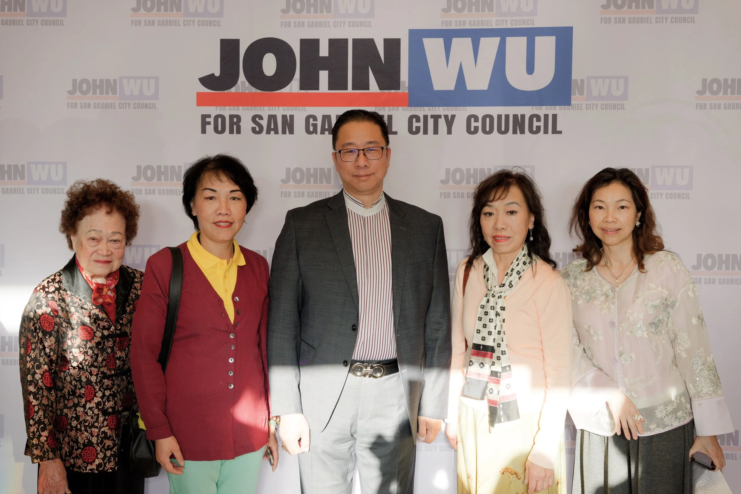 Group of six people standing in front of a campaign banner for John Wu running for San Gabriel City Council. The background features repeated text that says 'JOHN WU FOR SAN GABRIEL CITY COUNCIL'.