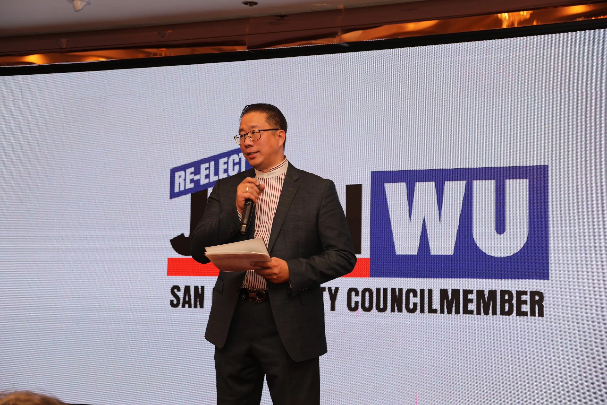 A man in a dark suit and glasses standing in front of a projection screen, holding a microphone in one hand and a stack of papers in the other. The screen displays the text "RE-ELECT" and "WILLI" with additional smaller text below.