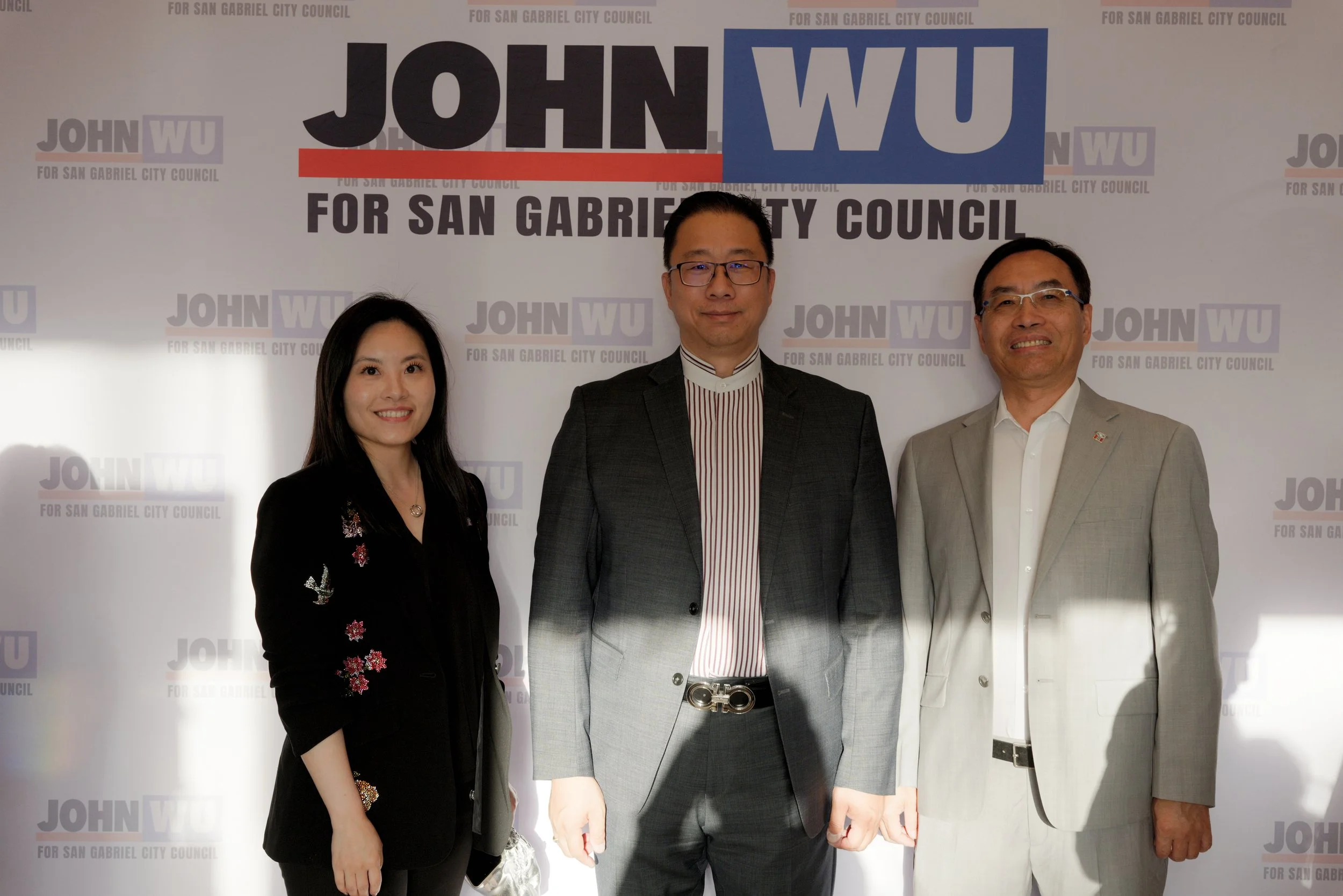 Group of three individuals standing in front of a backdrop with campaign text and logos. The backdrop reads 'JOHN WU FOR SAN GABRIEL CITY COUNCIL' repeatedly. The woman on the left is wearing a black blazer with floral embroidery, the man in the midd