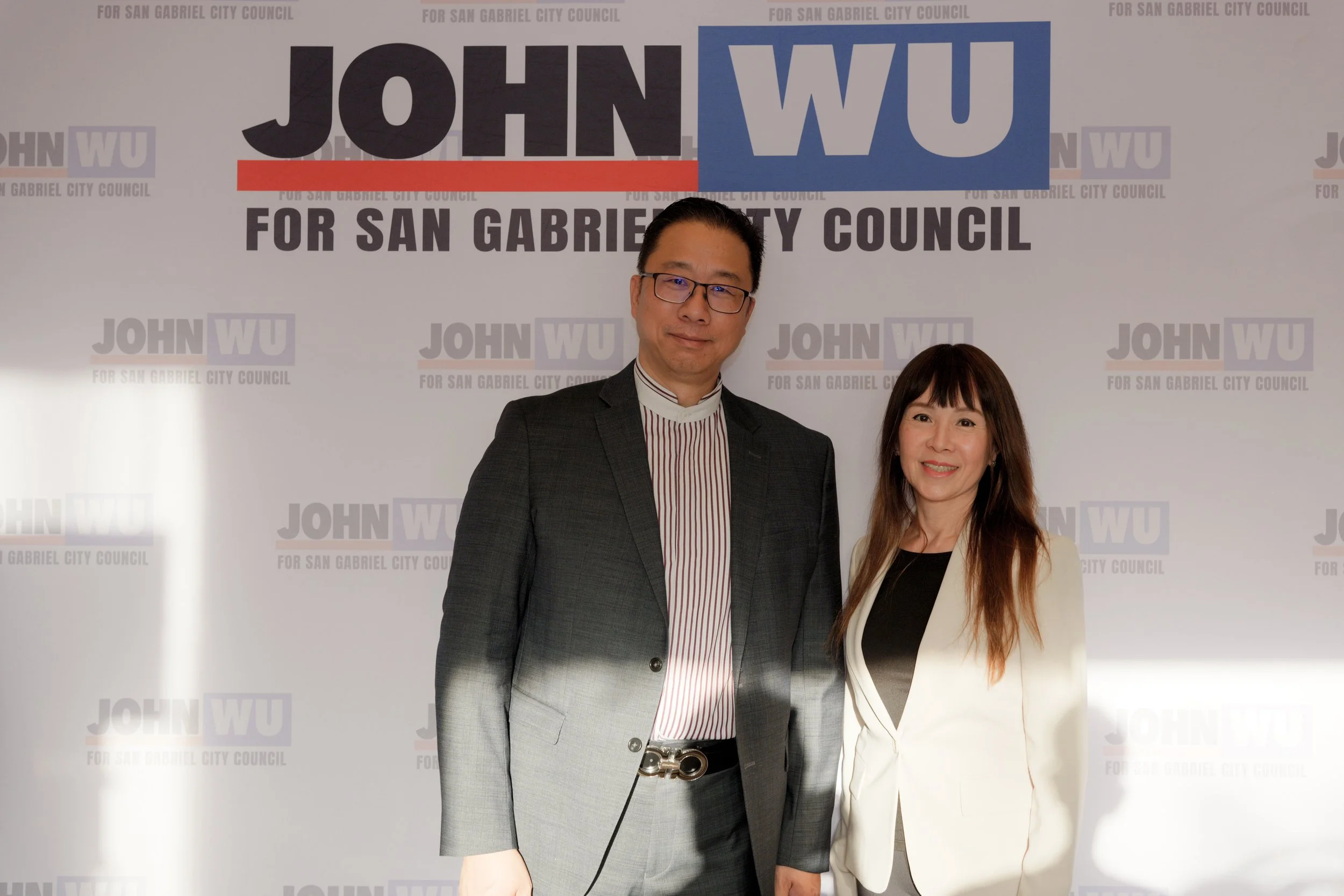 A man and a woman standing side by side in front of a campaign backdrop for John Wu running for San Gabriel City Council. The man is wearing glasses, a dark suit, and a striped shirt, while the woman is wearing a white blazer and black top. They are 