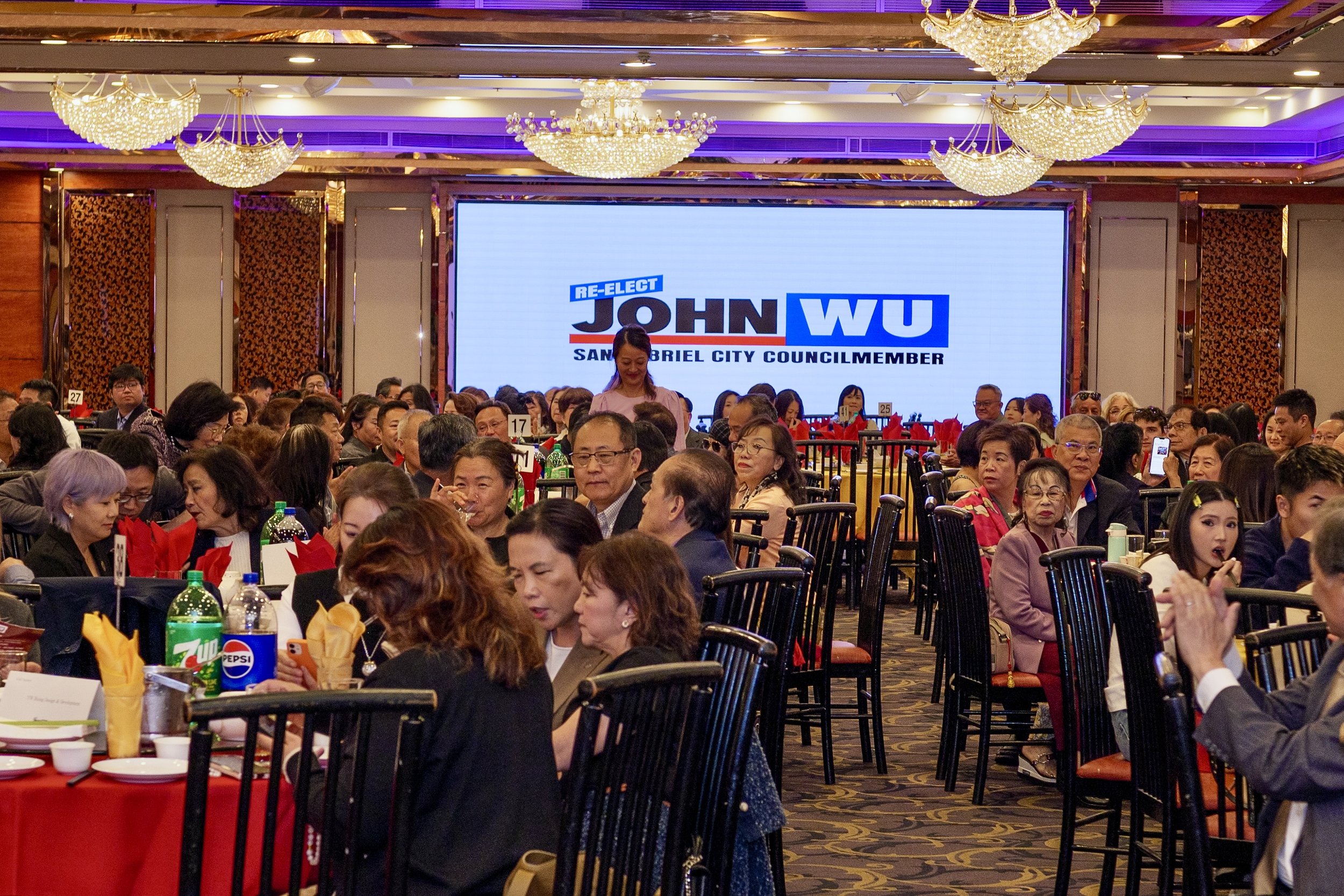 A large banquet hall filled with many people seated at round tables, with a big screen behind displaying a campaign sign for John Wu running for San Gabriel City Council. The hall is decorated with ornate chandeliers and patterned walls.