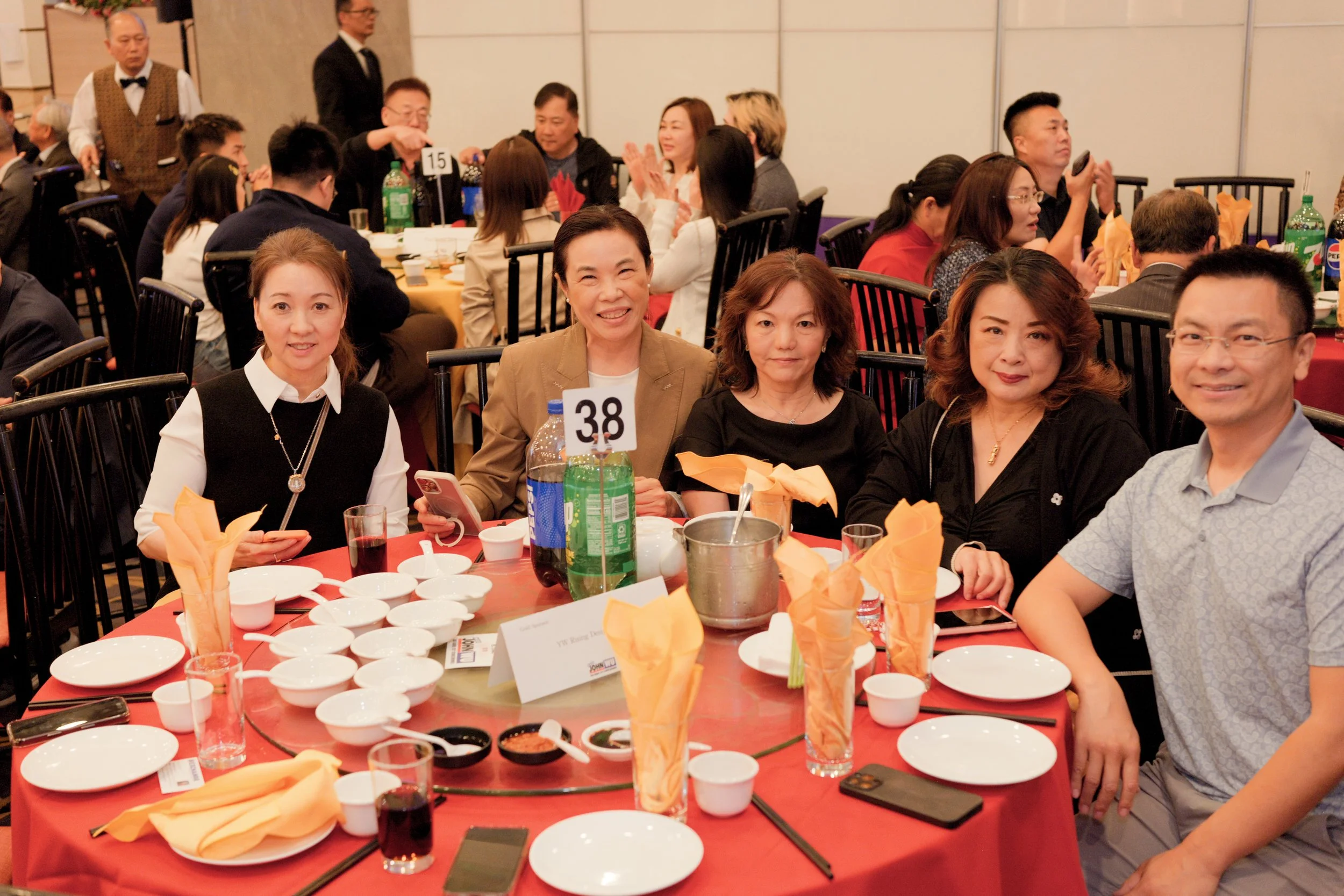 People sitting at round tables during a banquet or celebration, with dishes, drinks, and orange napkins on the table.