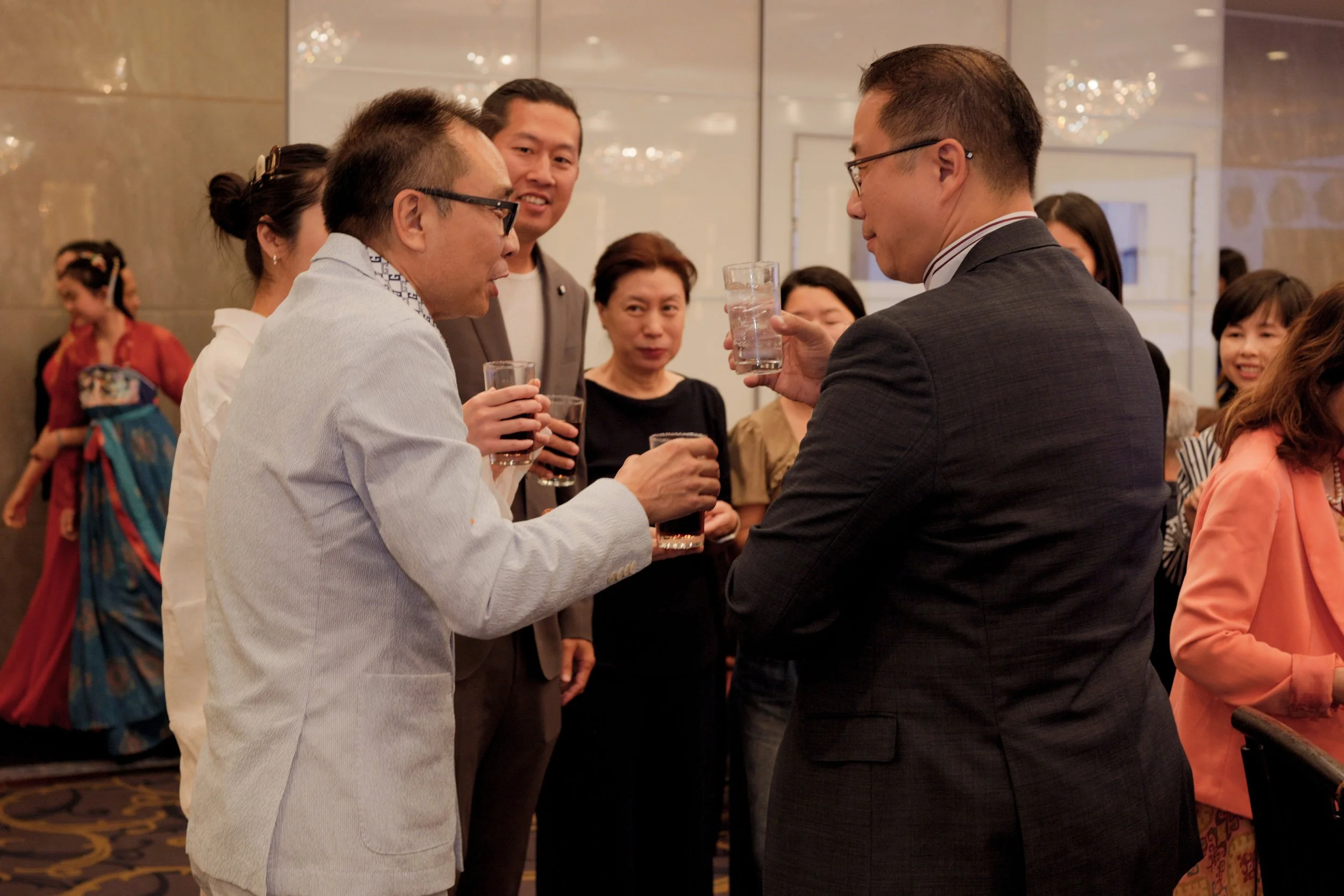 A group of people at a social gathering, with two men in suits and glasses in the center toasting with clear glasses of water, while others watch and smile in a well-lit, elegant room with chandeliers.