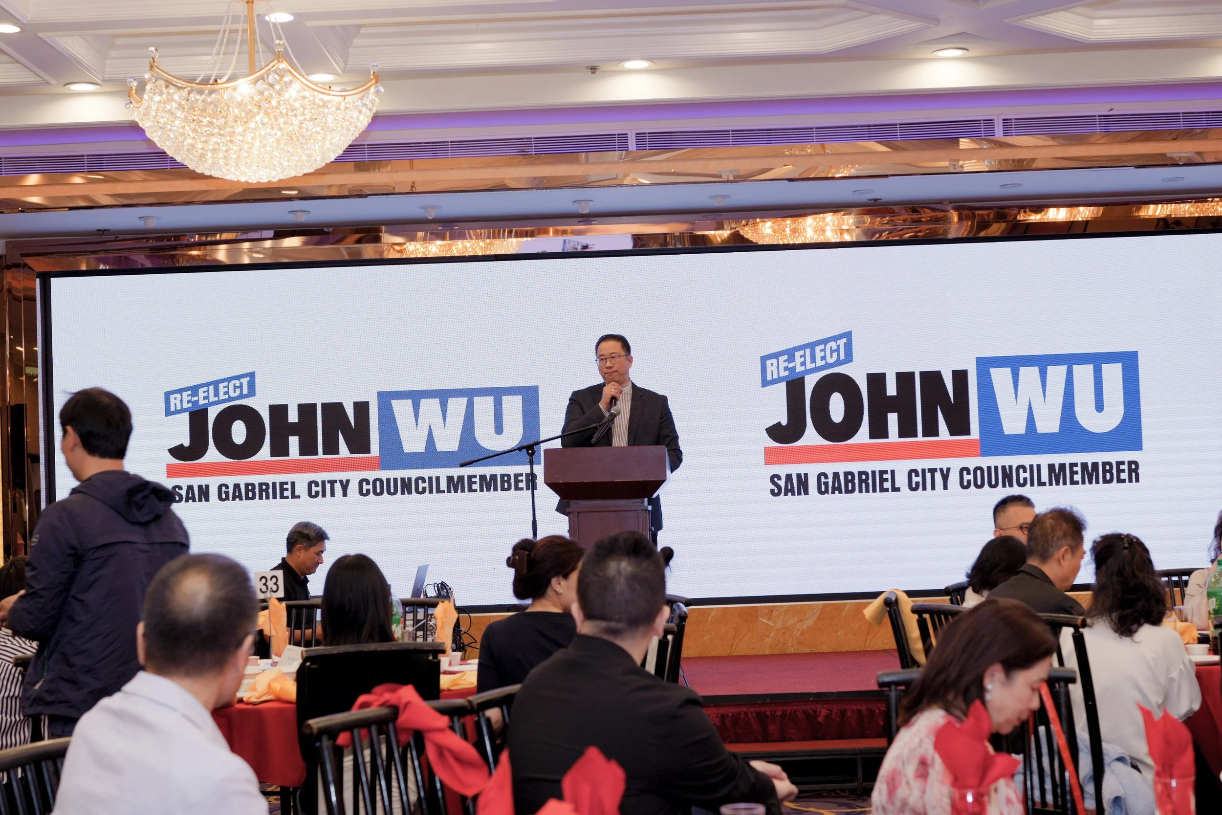 A man stands at a podium speaking at a political event with a large screen behind him showing the words "Re-elect John Wu, San Gabriel City Councilmember." There are many people sitting at tables and listening, with some wearing red clothing, in an e