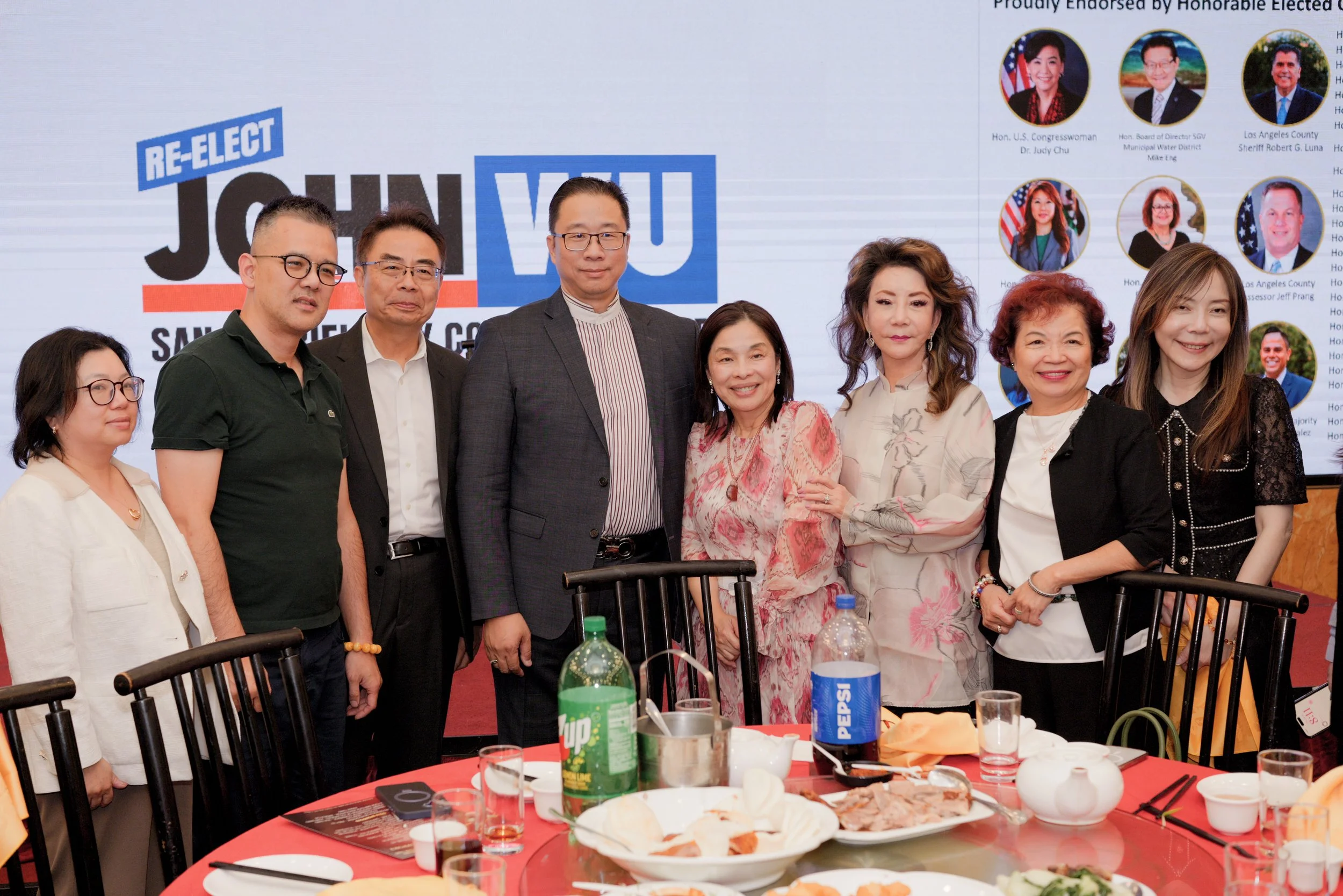 Group of seven people standing behind a table at a celebratory event, with food and beverages, in front of a large screen displaying 'Re-Elect Judy'.