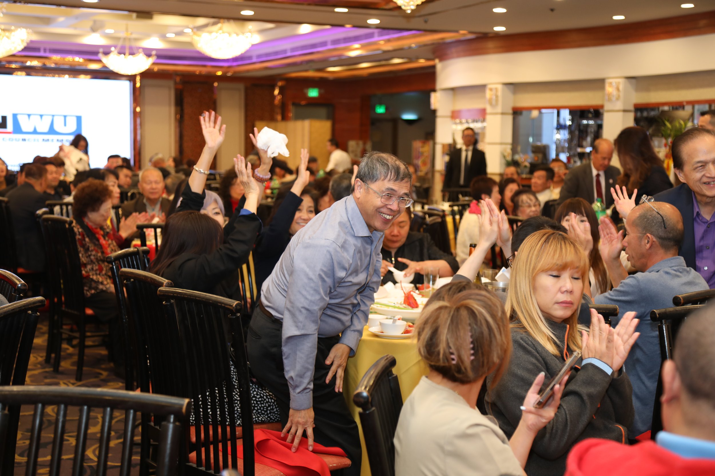 People attending a banquet or celebration, seated at tables, some raising hands, with a brightly lit and decorated banquet hall in the background.