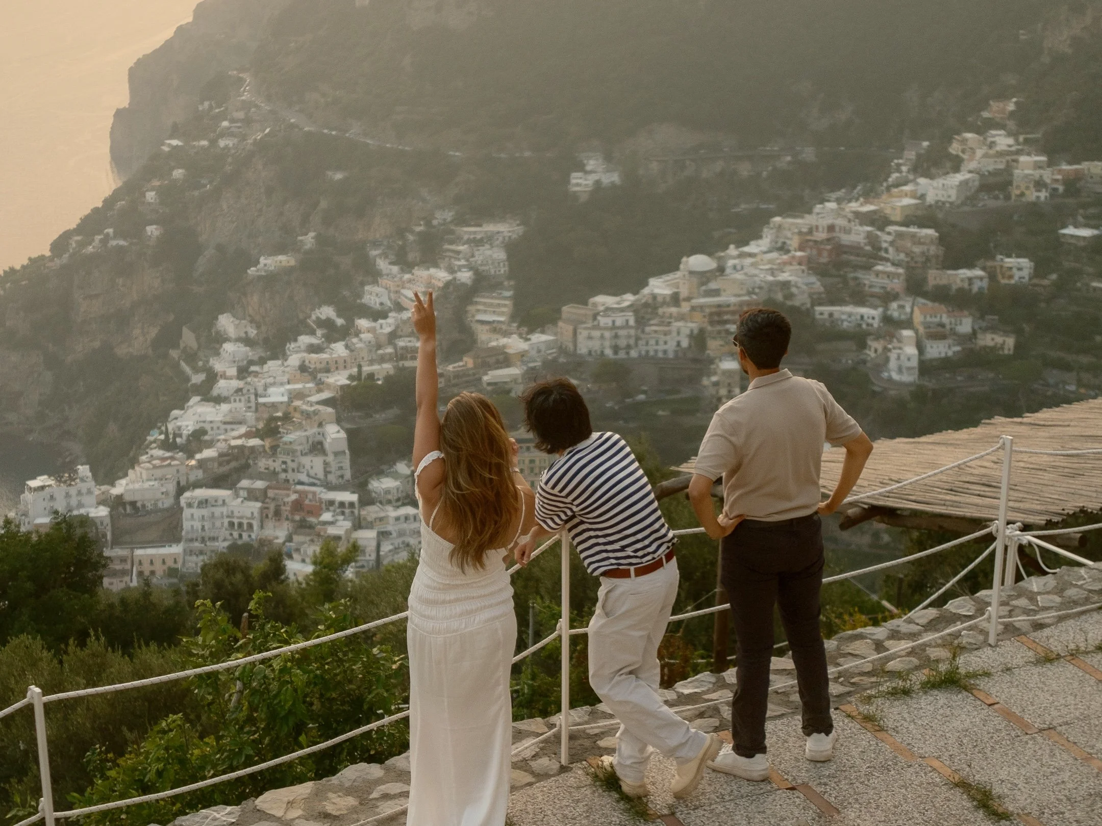 Group of four people overlooking a hillside town, one woman raising her hand with a peace sign, all facing away from the camera at sunset.
