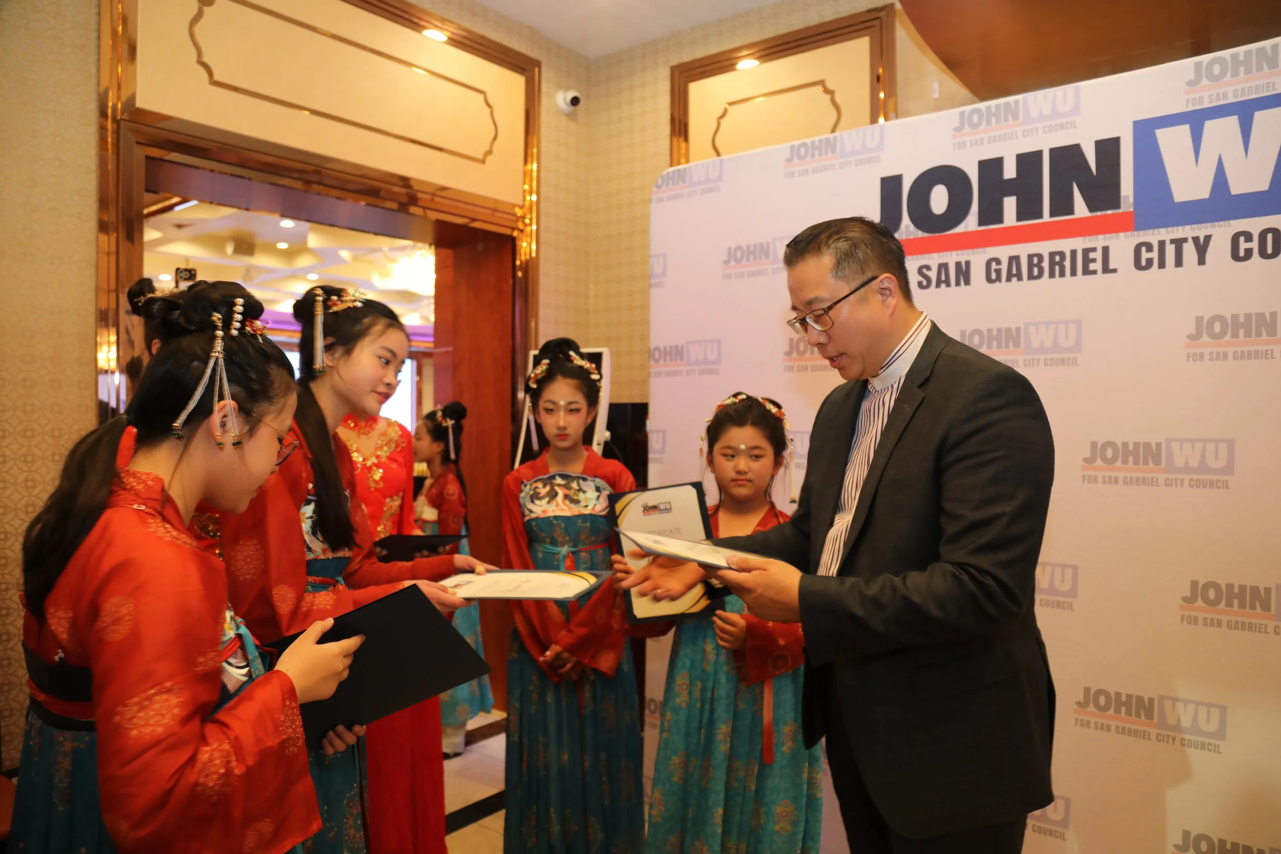 A group of girls dressed in traditional Chinese attire receiving diplomas or certificates from a man in a suit at an event promoting John Wu for San Gabriel City Council.