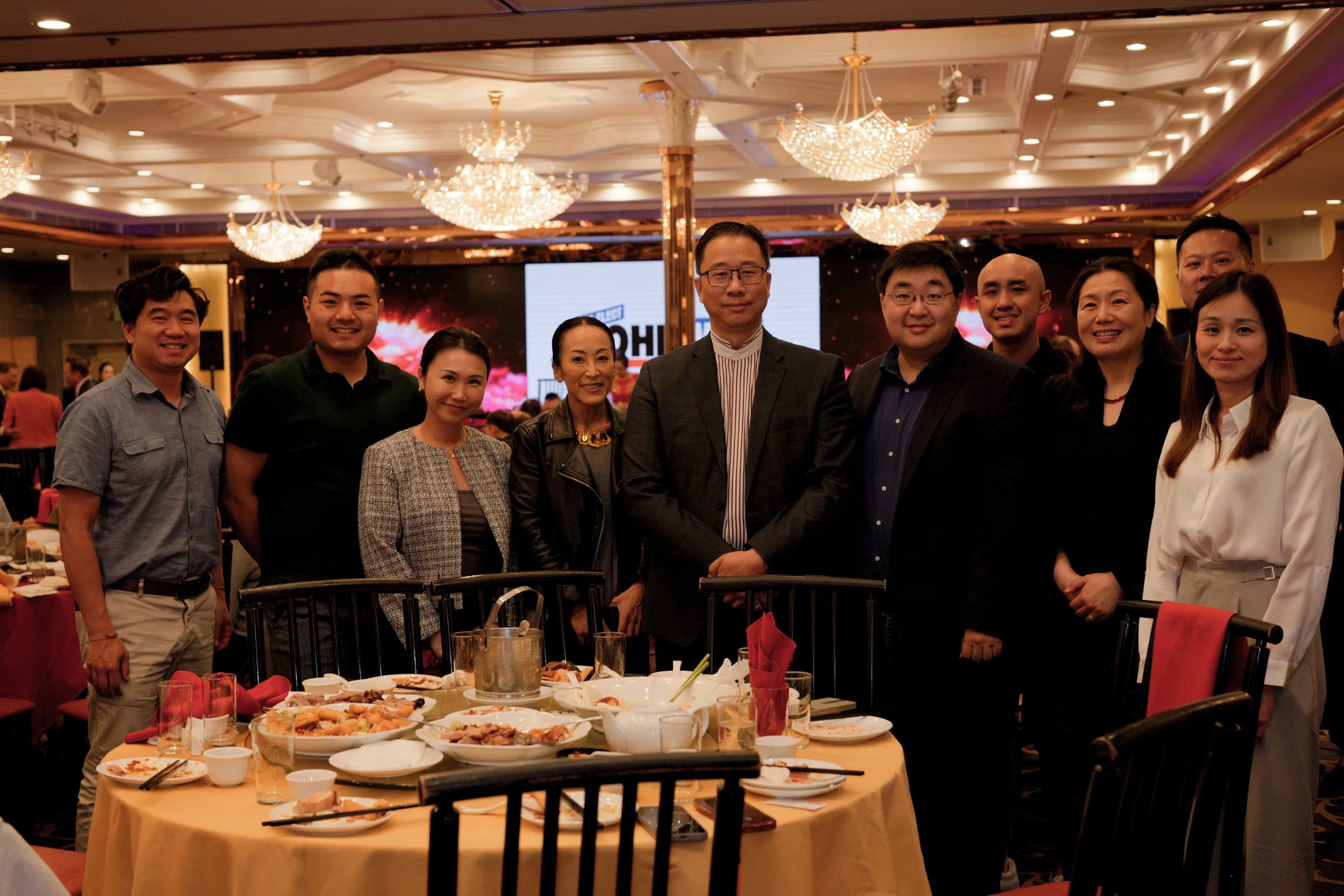 Group of people posing together at a dining event in a decorated banquet hall with chandeliers and a large digital screen in the background.