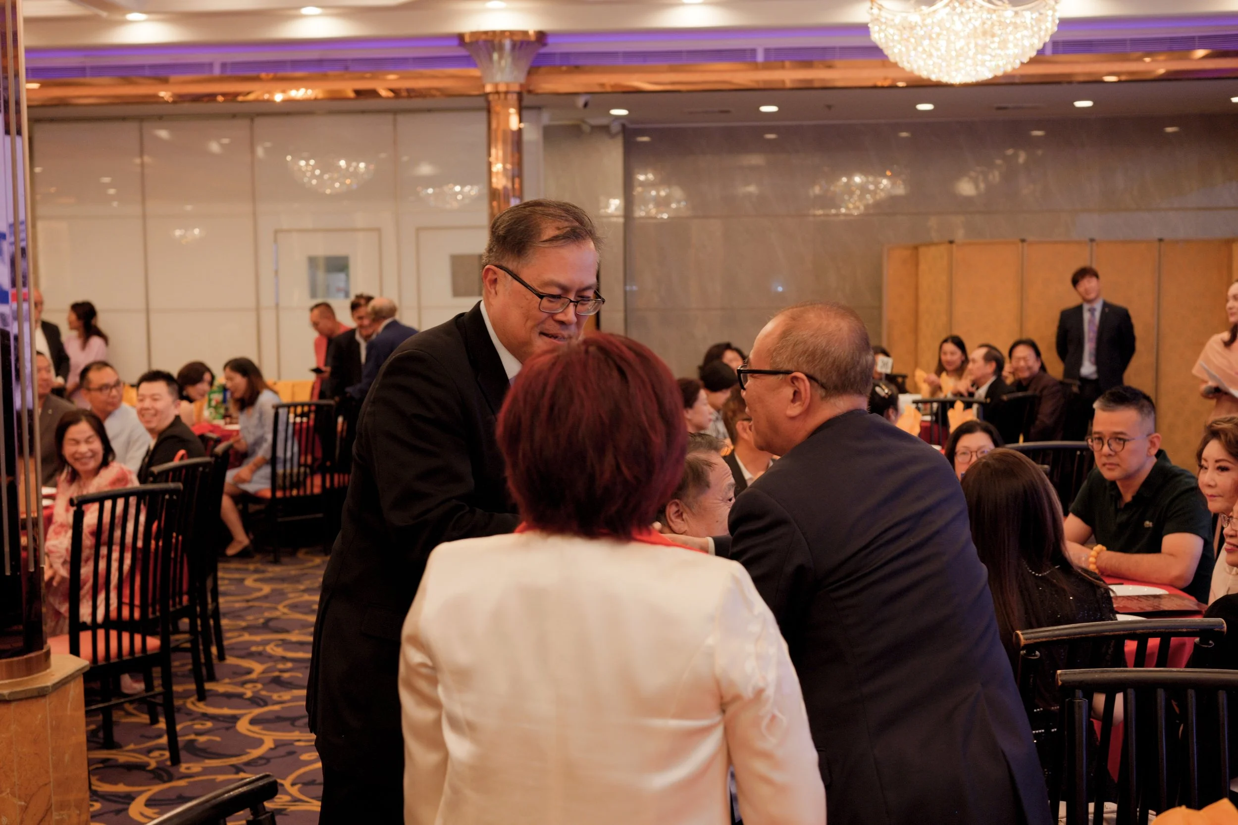 A large banquet hall filled with people sitting and socializing at round tables during a formal event. A man in a black suit and glasses is talking to a woman with red hair, who is facing away from the camera. The room is decorated with chandeliers a