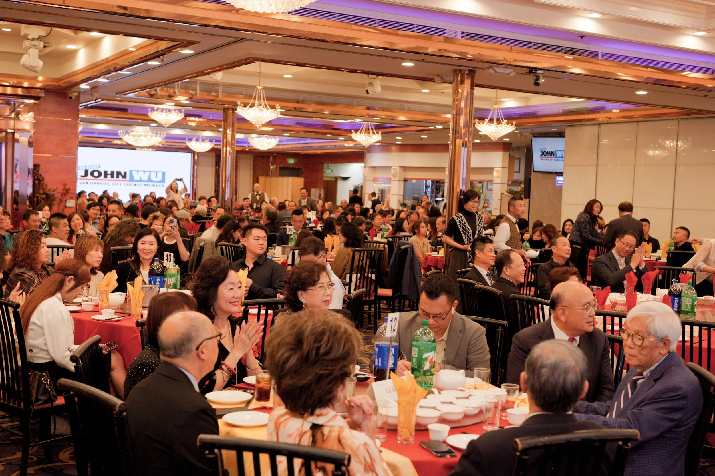 Crowded banquet hall with many people seated at round tables, enjoying a celebration or event, with chandeliers hanging from the ceiling and a large screen displaying campaign signs in the background.