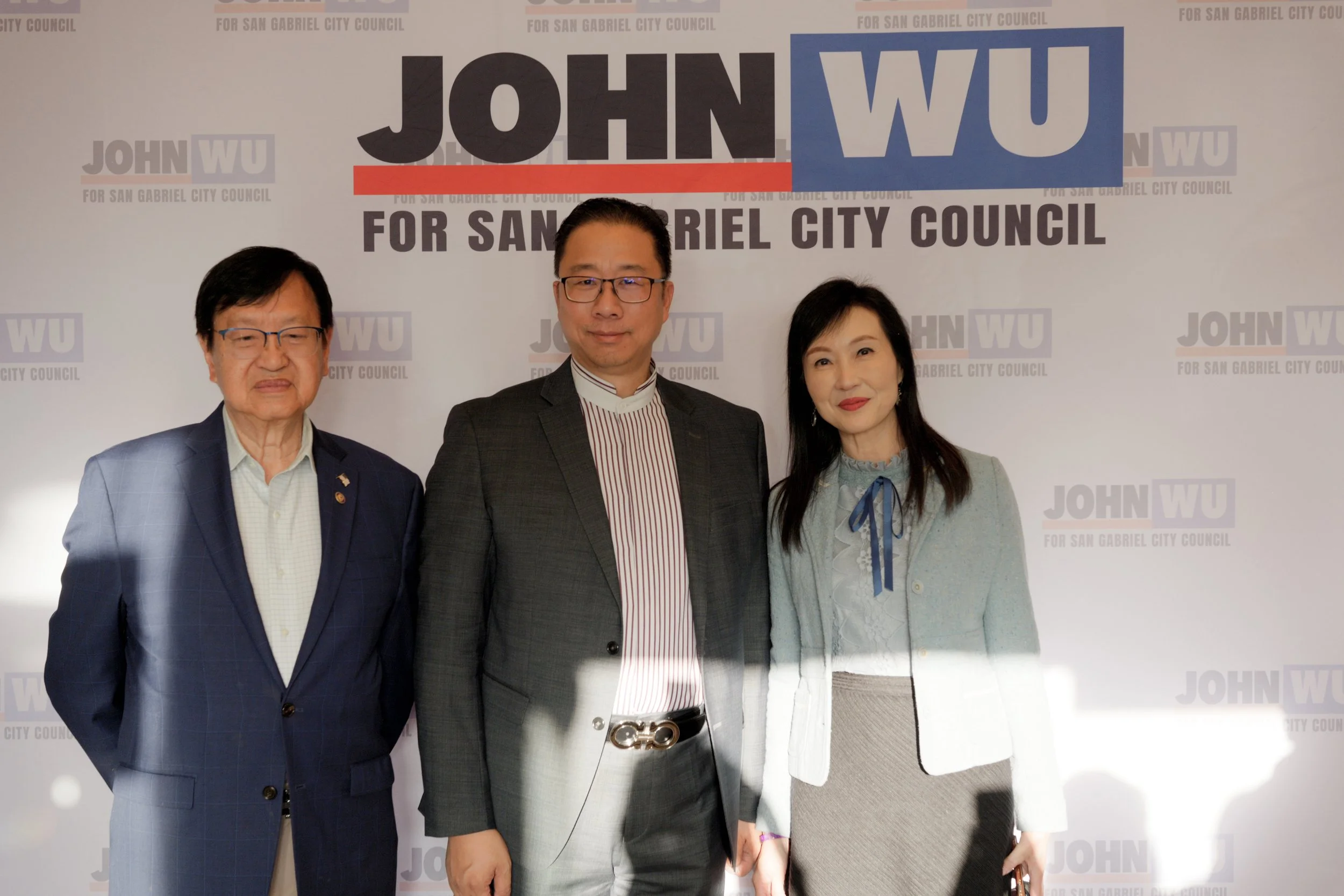 Three people standing in front of a backdrop with the text 'JOHN WU FOR SAN GABRIEL CITY COUNCIL' and multiple logos. They are dressed in formal attire and are posing for a photo.