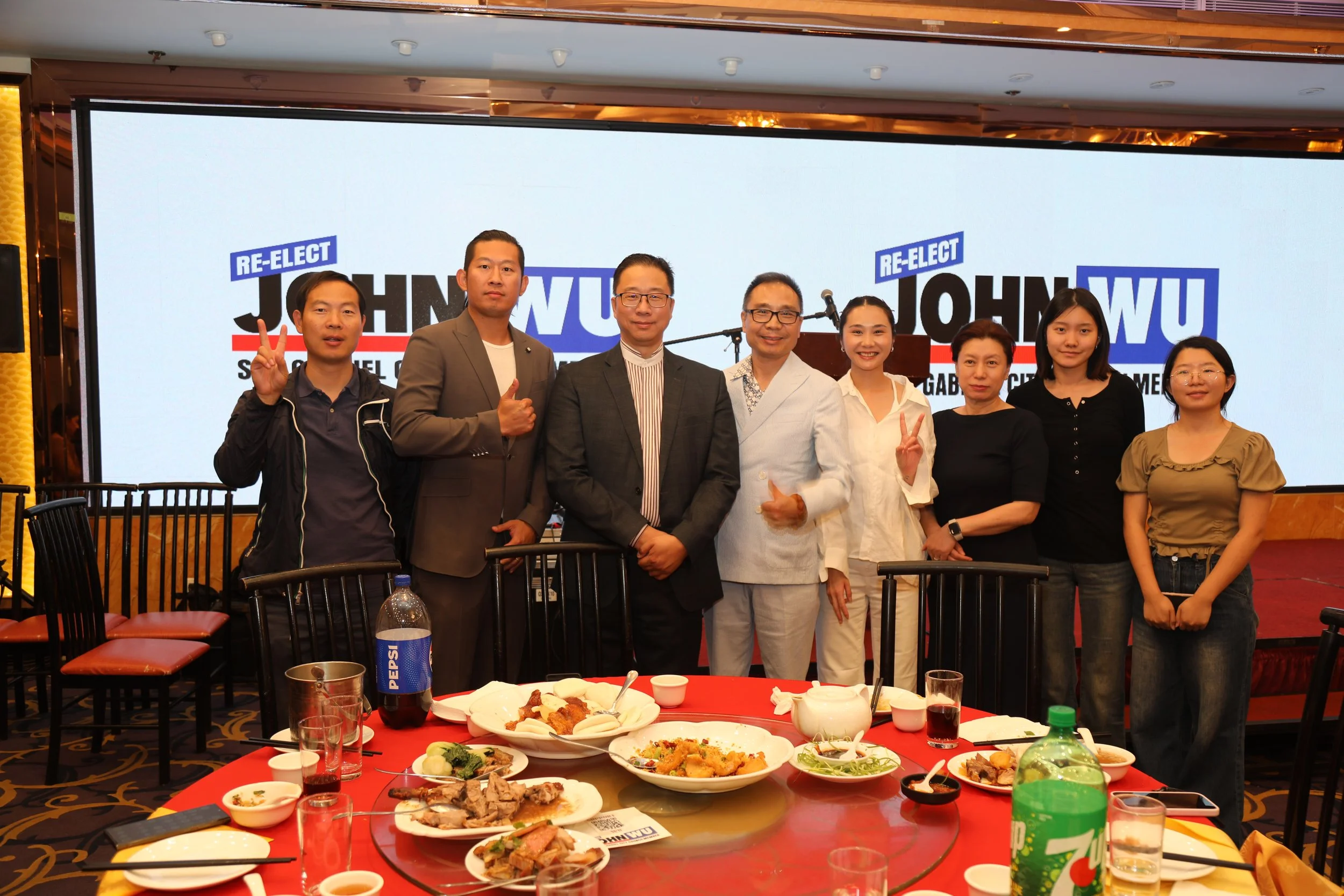Group of eight people standing together at a banquet table with food, in front of a screen displaying the text 'Re-elect John Wu'.