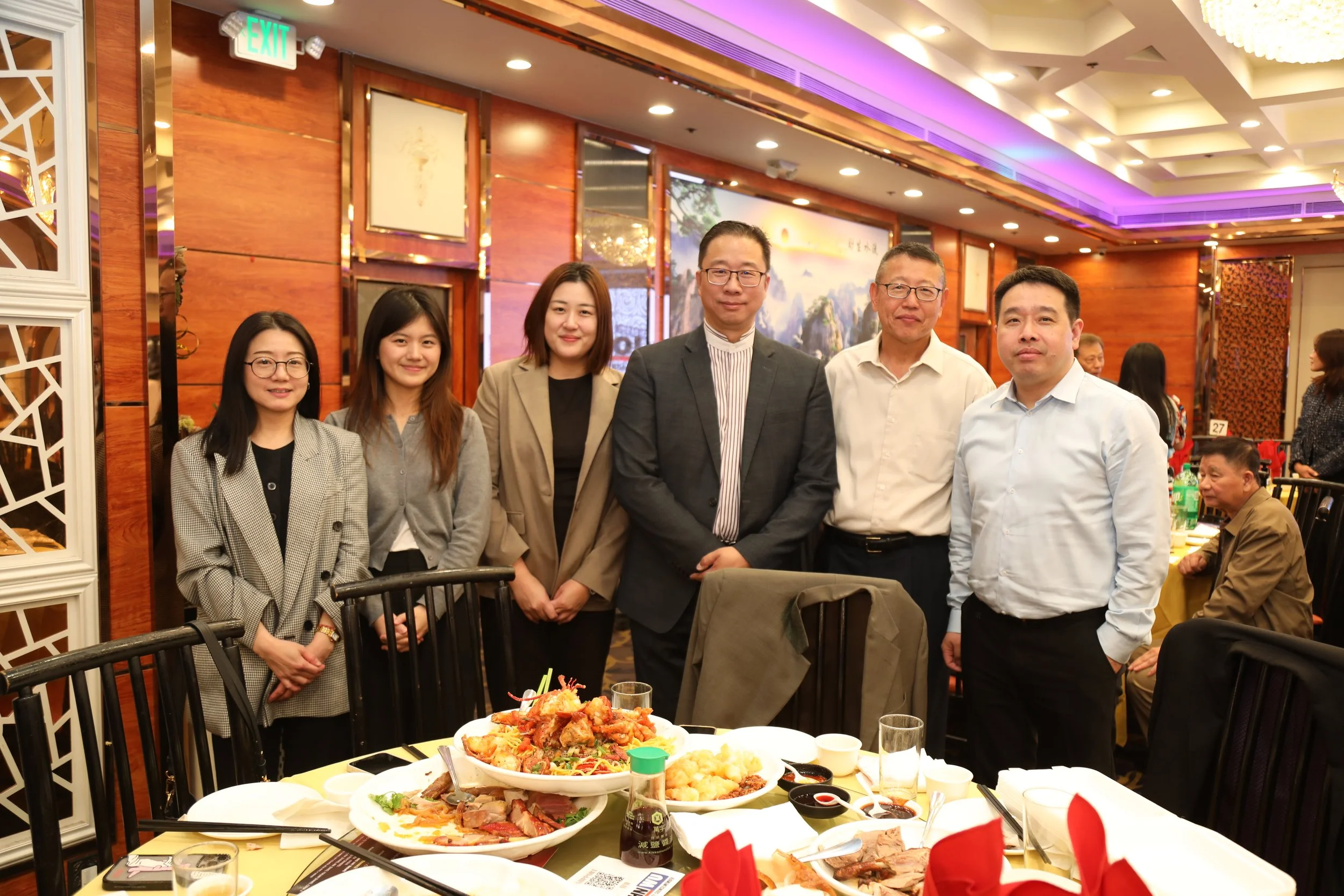 Group of six people standing together at a restaurant dinner, with food on the table in front of them, in a decorated dining room.