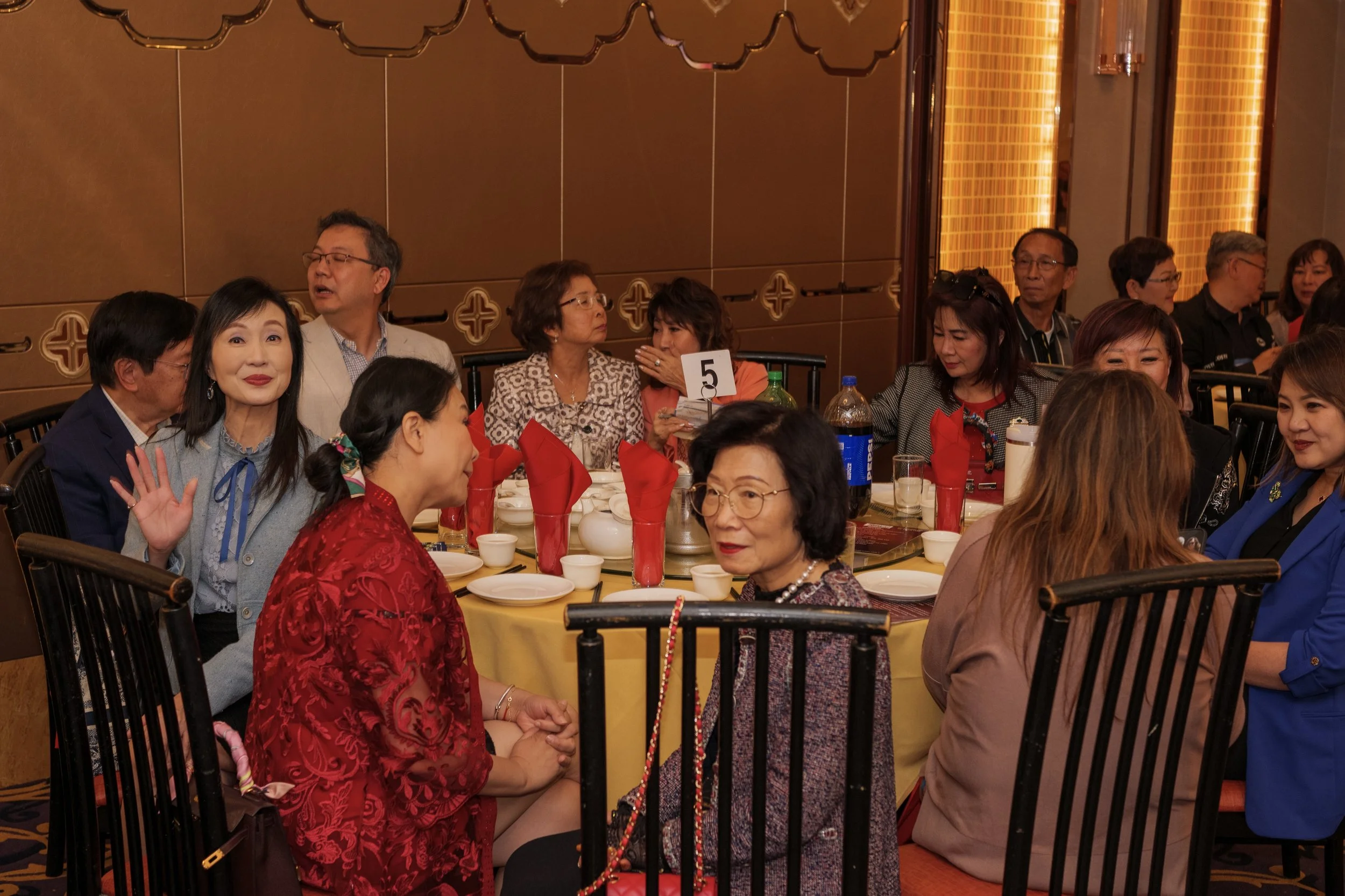 Group of Asian and Asian American women and men seated around a banquet table at a social or celebratory event, with red napkins, bottles of soda and small bowls.
