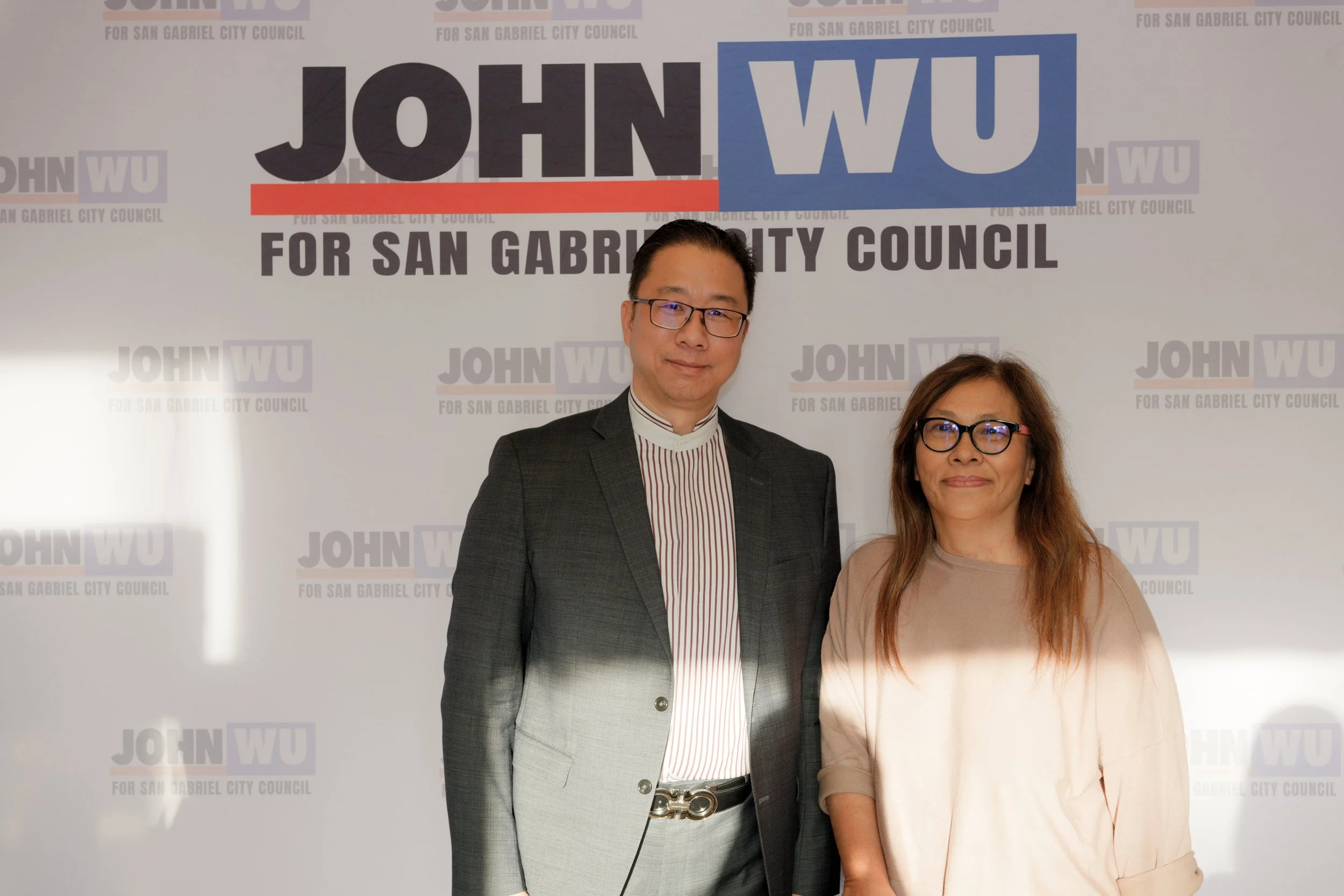 Two people standing in front of a background with campaign slogans and logos for San Gabriel City Council. The man on the left is wearing glasses, a dark blazer, and a striped shirt. The woman on the right has long hair, glasses, and is wearing a lig