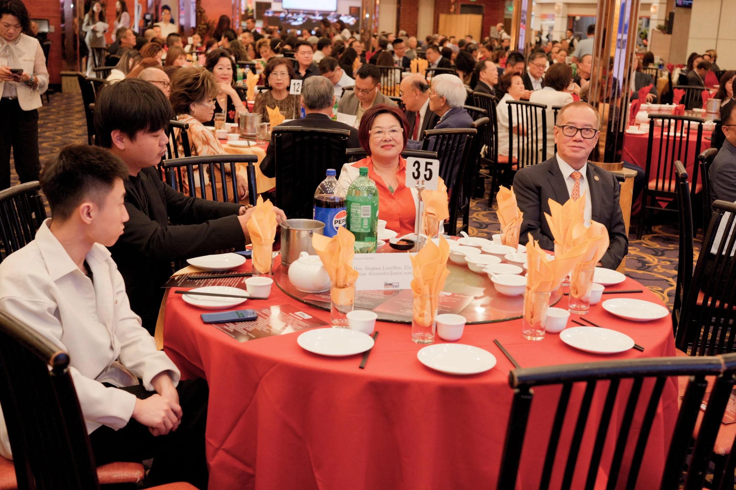 People attending a banquet in a large indoor venue with tables set for a meal, decorated with orange napkins, bottles of soda, and dinnerware, with many dressed in formal attire.