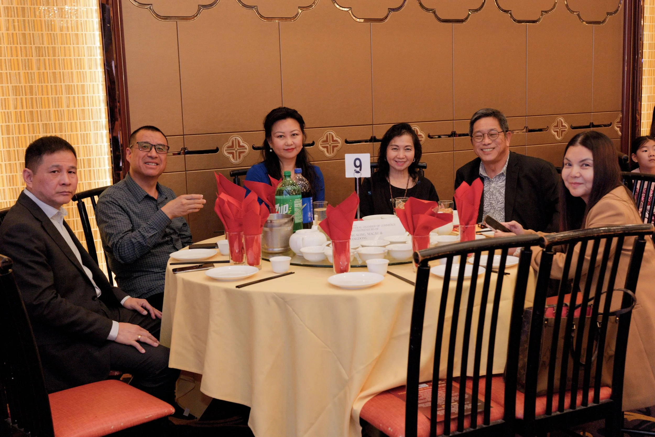 A group of seven people, five women and two men, sitting around a banquet table at a restaurant during a formal event. The table is decorated with red napkins, beverages, and white dishes. The background features a brown partition with decorative pat