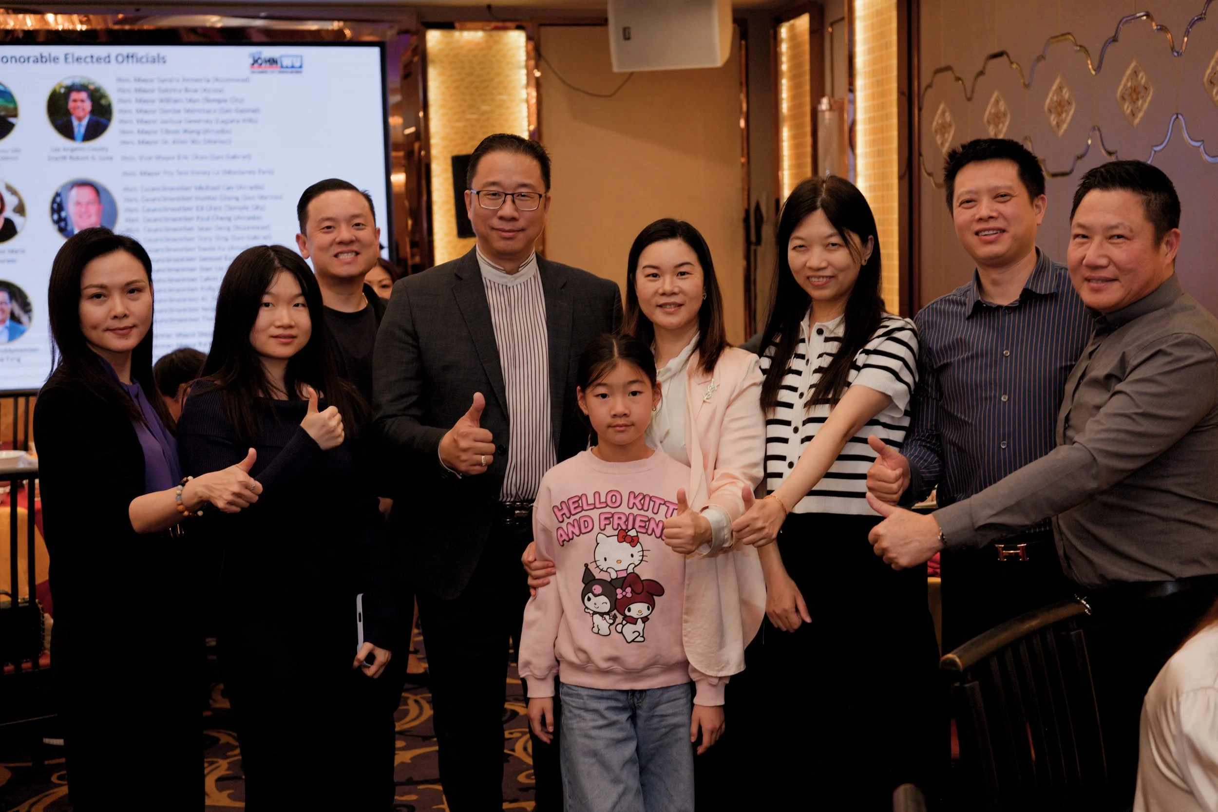 Group of nine people, including children, standing together indoors at a formal or celebratory event, with a large screen displaying information about elected officials in the background.